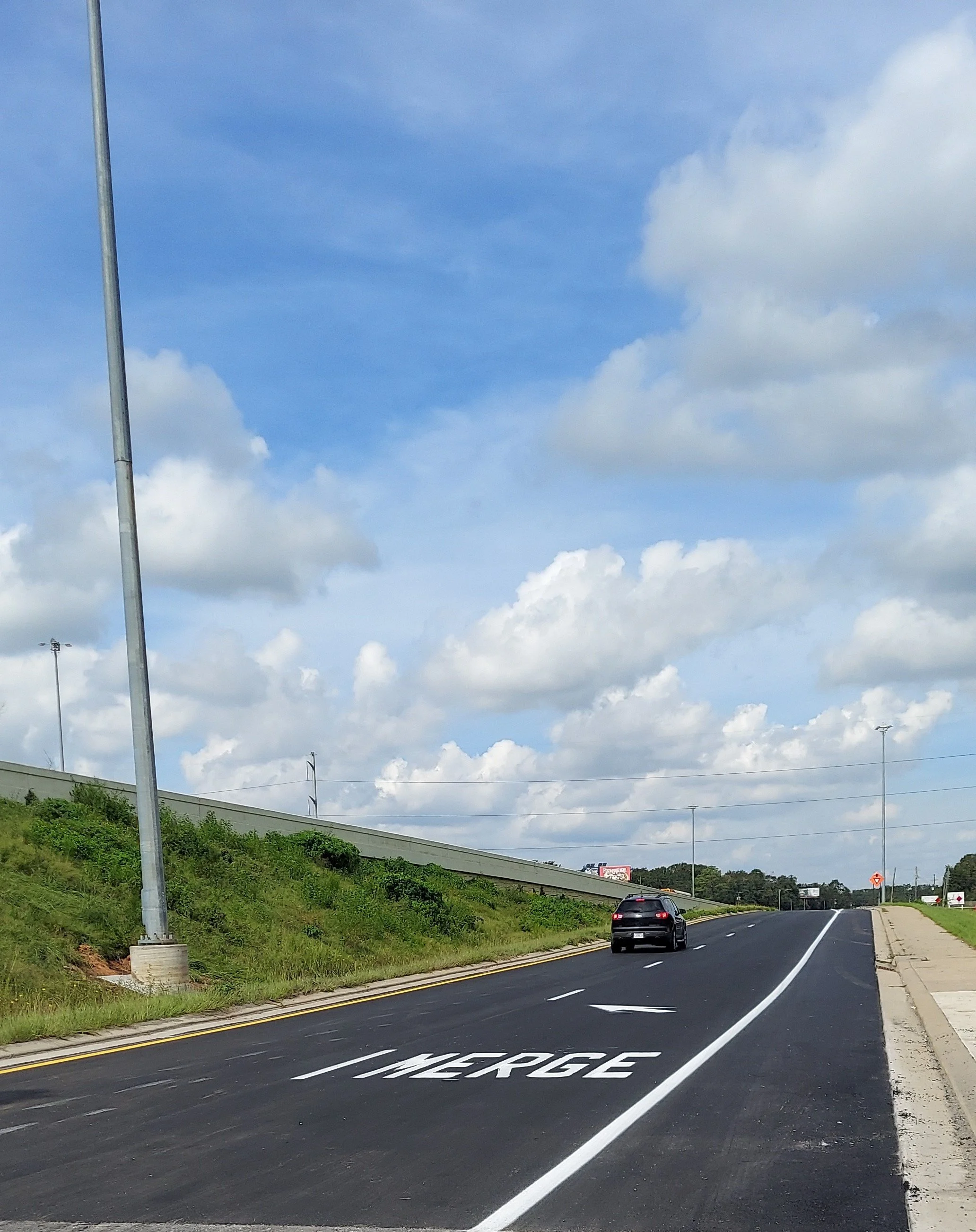 An empty roadway with a black car driving on it, a sign that says 'MERGE,' a grassy hill on the left, cloudy sky above.