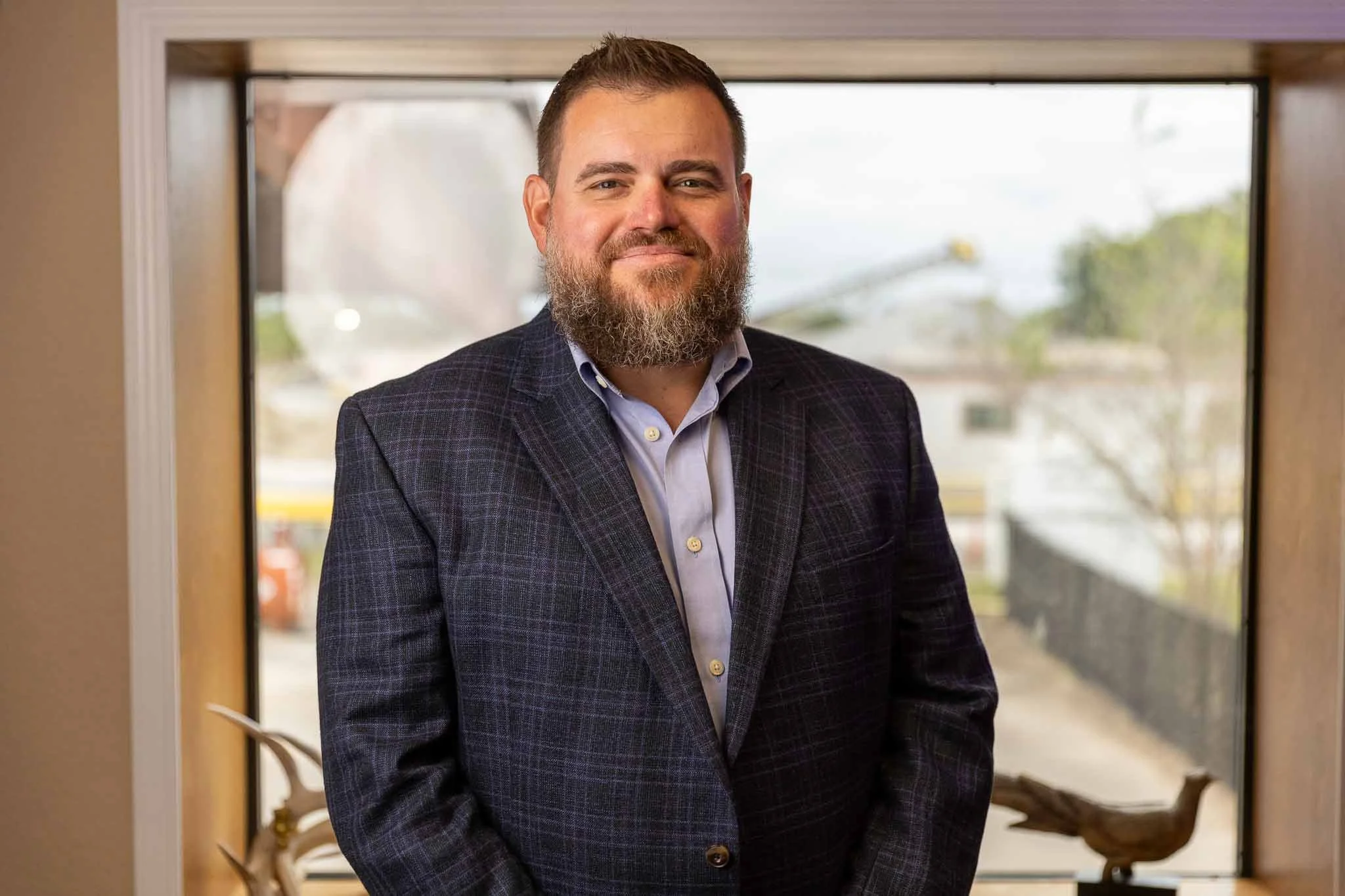 Man with a beard and short hair wearing a dark checkered suit jacket and light blue dress shirt standing indoors in front of a large window with a blurred outdoor background.