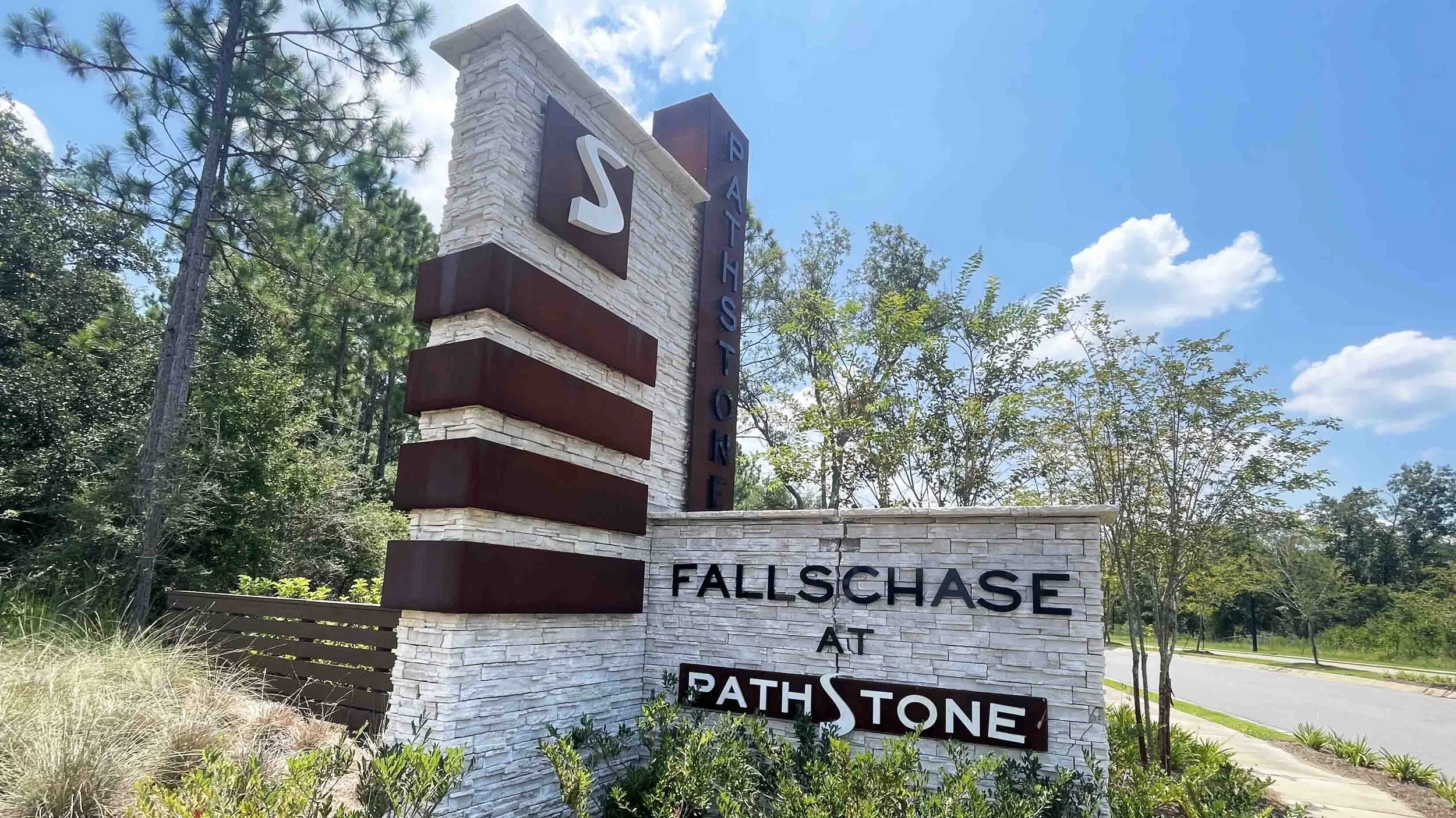 A sign for the community of Fallschase at Pathstone, featuring a modern stone and metal design with the community's name and logo, set against a backdrop of trees and a blue sky.