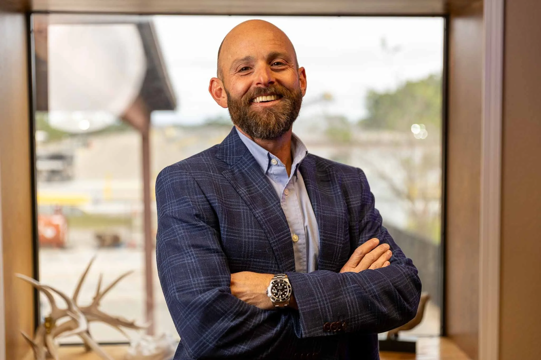 A smiling bald man with a beard, wearing a blue checkered blazer, light blue shirt, and wristwatch, standing with arms crossed in front of a large window with a blurred outdoor background.