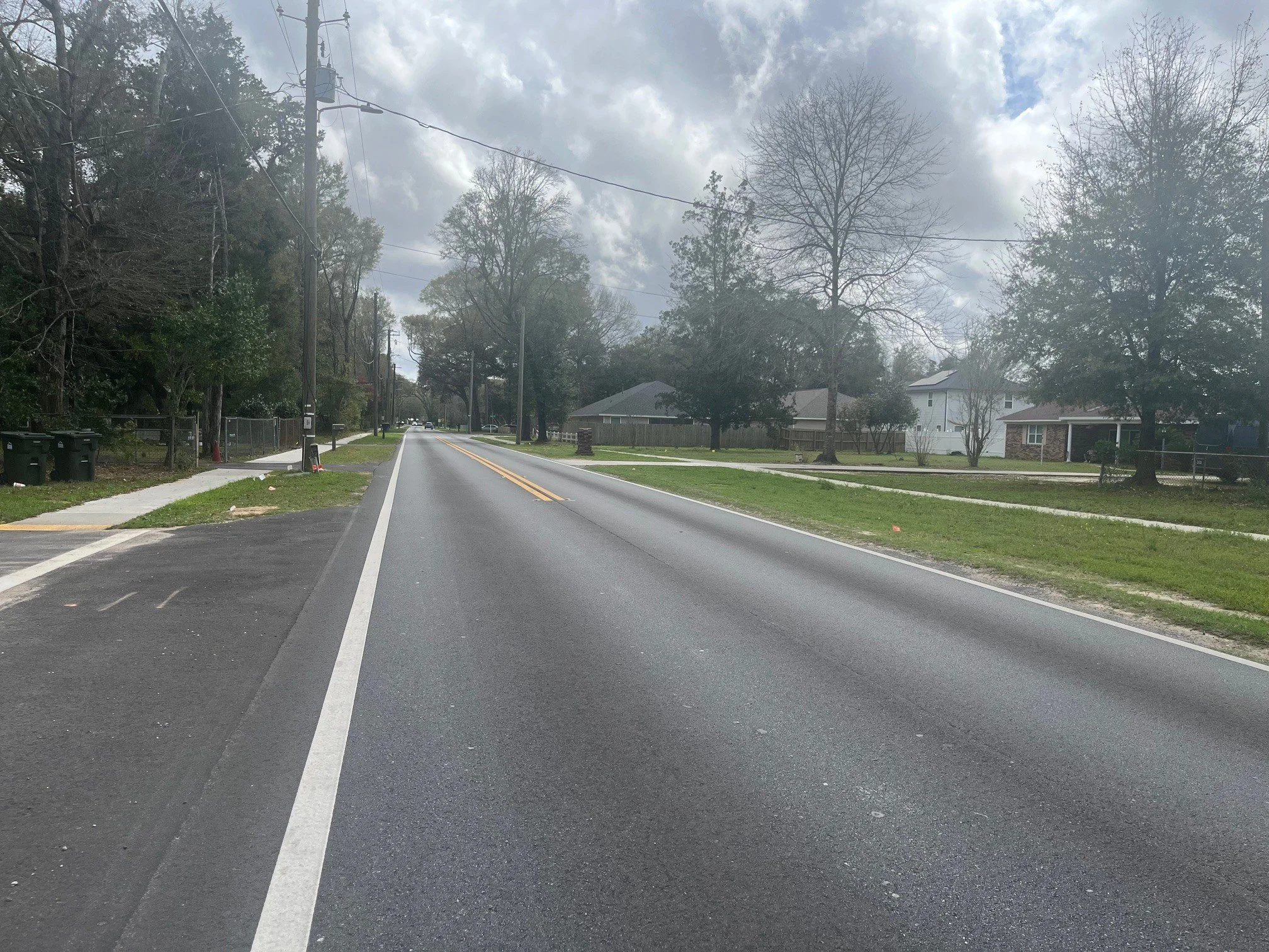 A quiet residential street with a two-lane road, houses on the right, and trees lining both sides. The sky is cloudy, and the street appears to be in a suburban area.