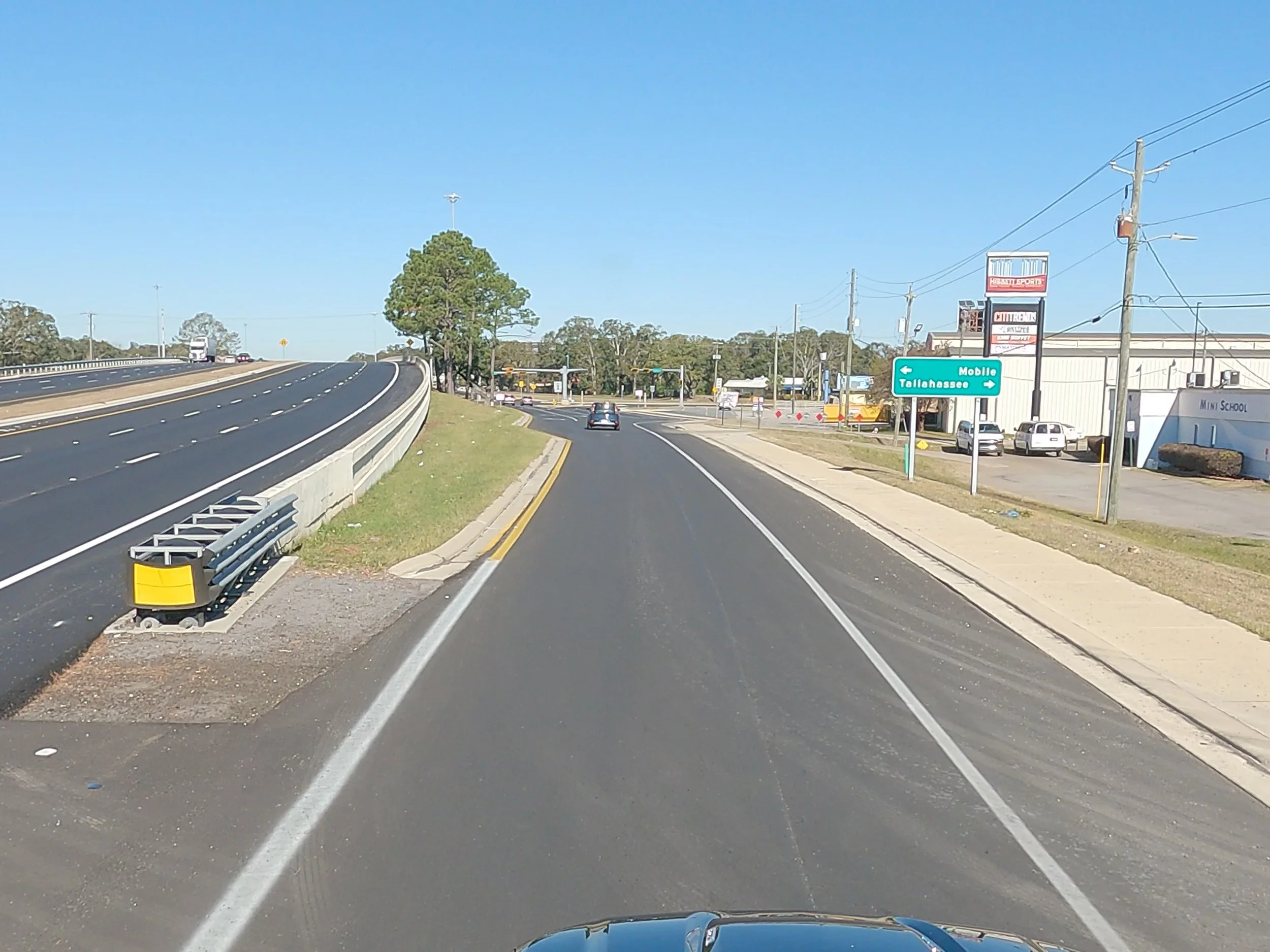 A two-lane road with a curved on ramp, a car driving ahead, and a blue directional sign for Mobile and Tallahassee on the right side. Utility poles and a building with a sign are visible on the right side of the road. The sky is clear and blue.