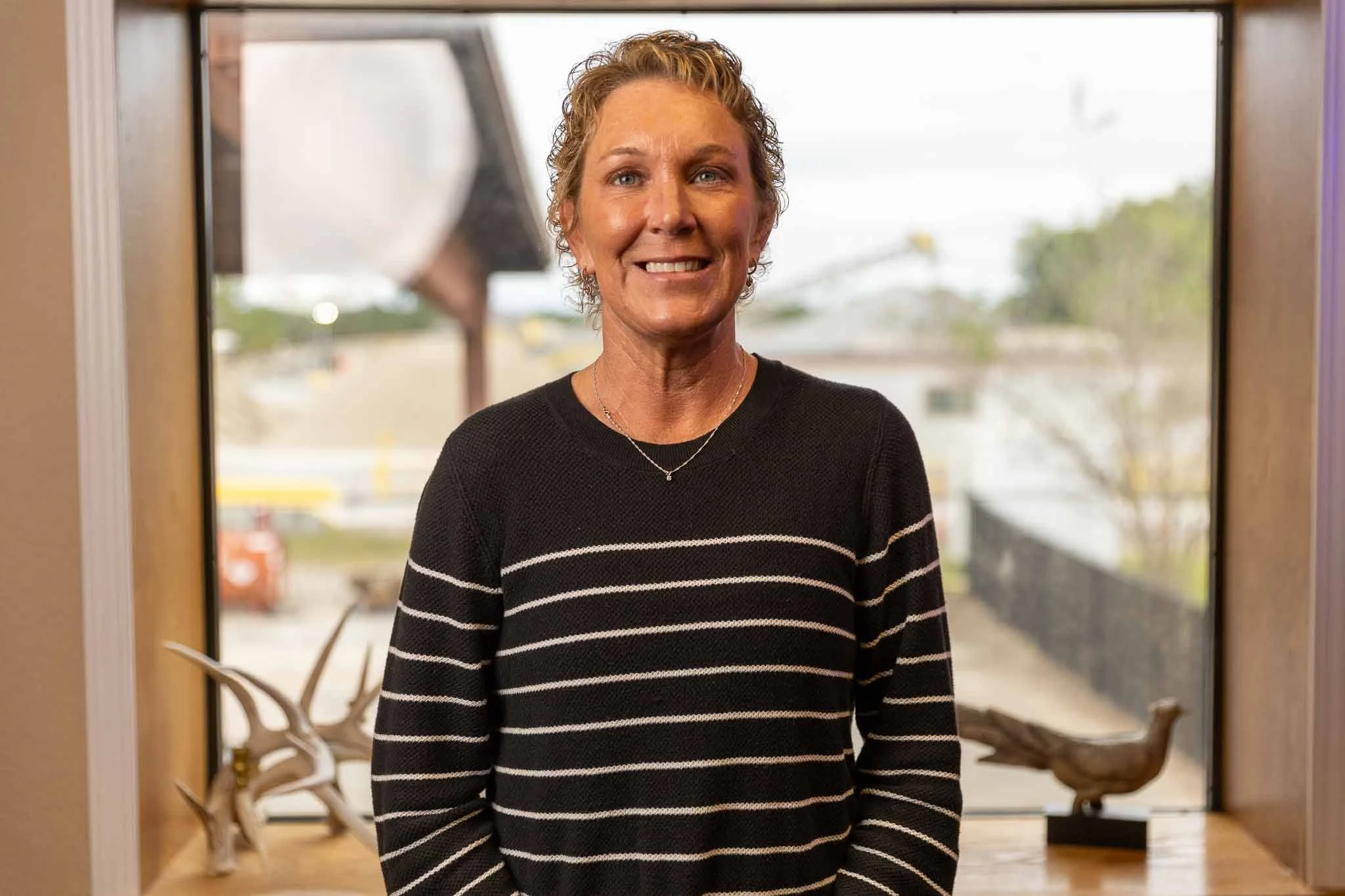 A smiling woman with curly blonde hair, wearing a black sweater with white horizontal stripes, standing indoors in front of a window with a blurred outdoor background.