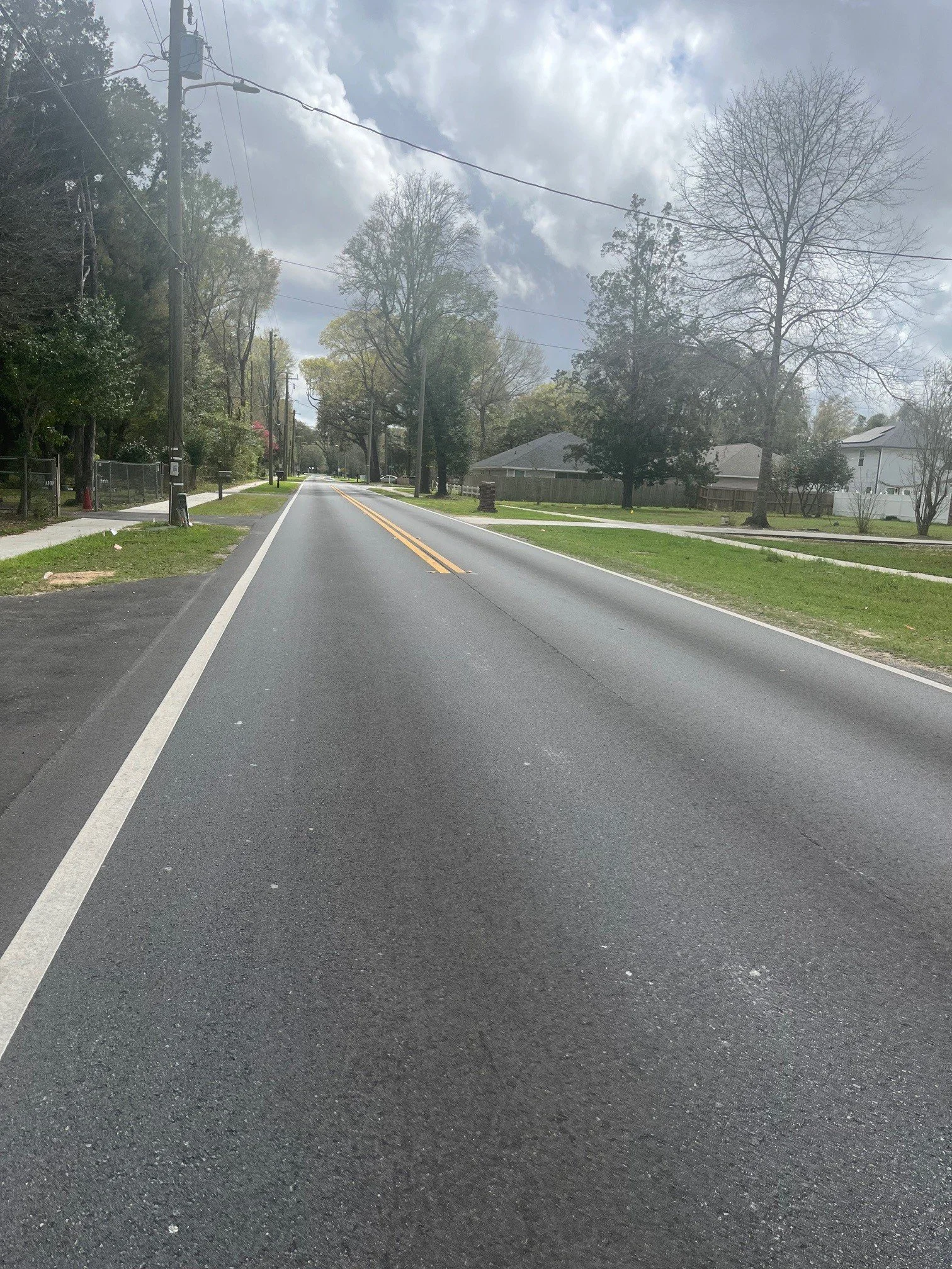 A quiet suburban street with trees lining both sides, houses behind fences, and a cloudy sky overhead.