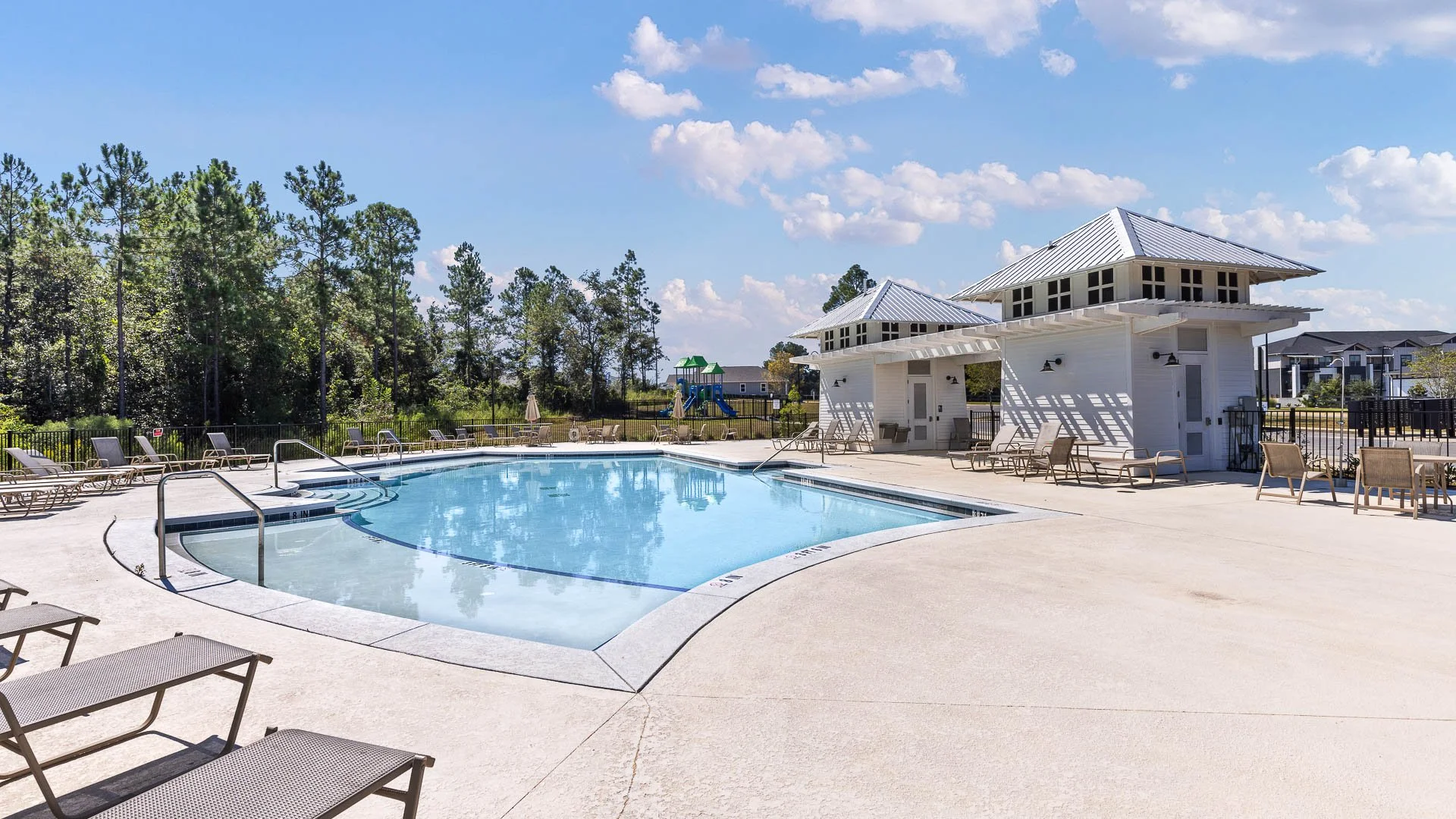 An outdoor swimming pool area with lounge chairs, a small building, and a playground with green slides in the background, surrounded by trees and a blue sky with some clouds.
