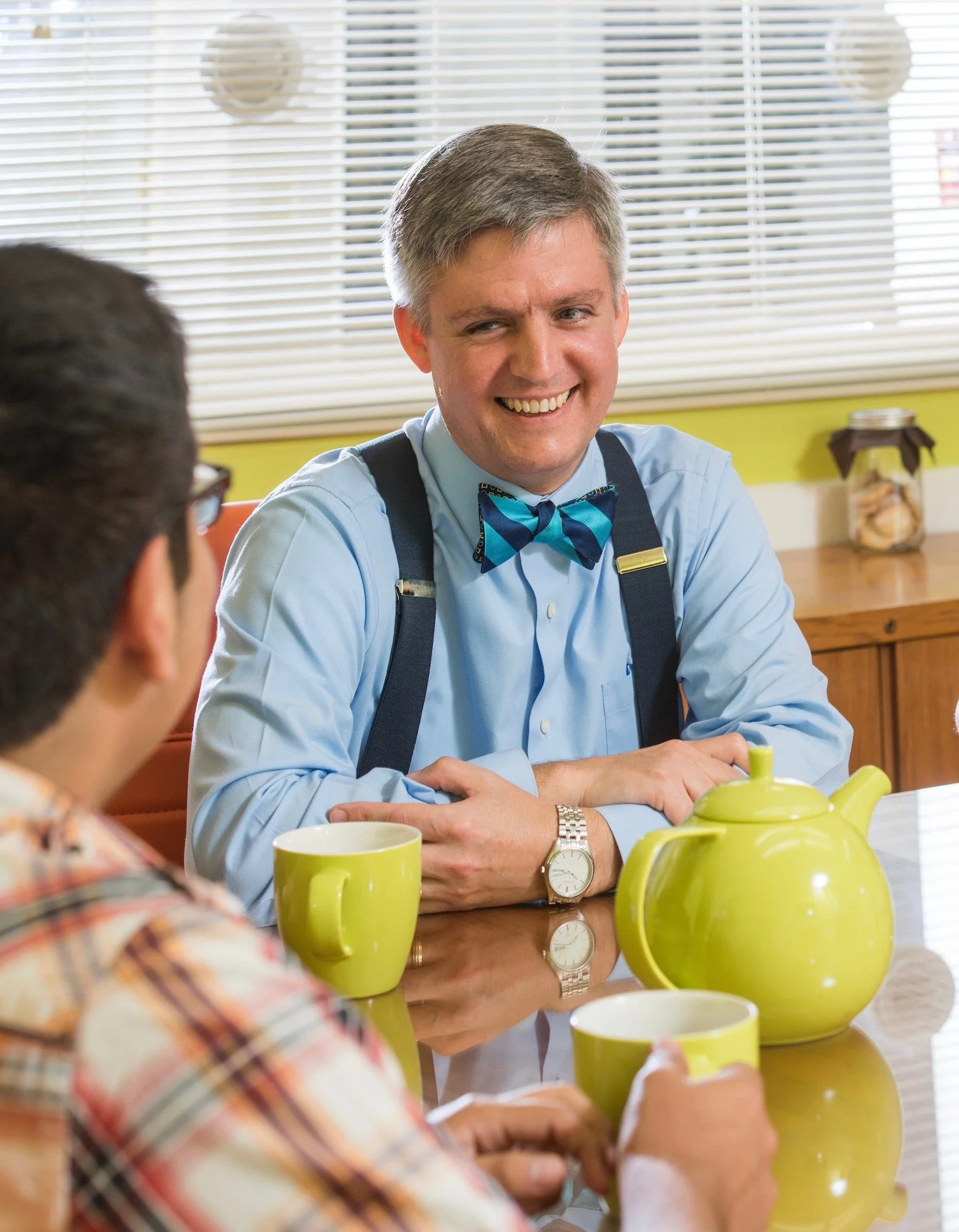 Two men sitting at a table with green coffee mugs and a green teapot, engaging in conversation, in a room with window blinds.