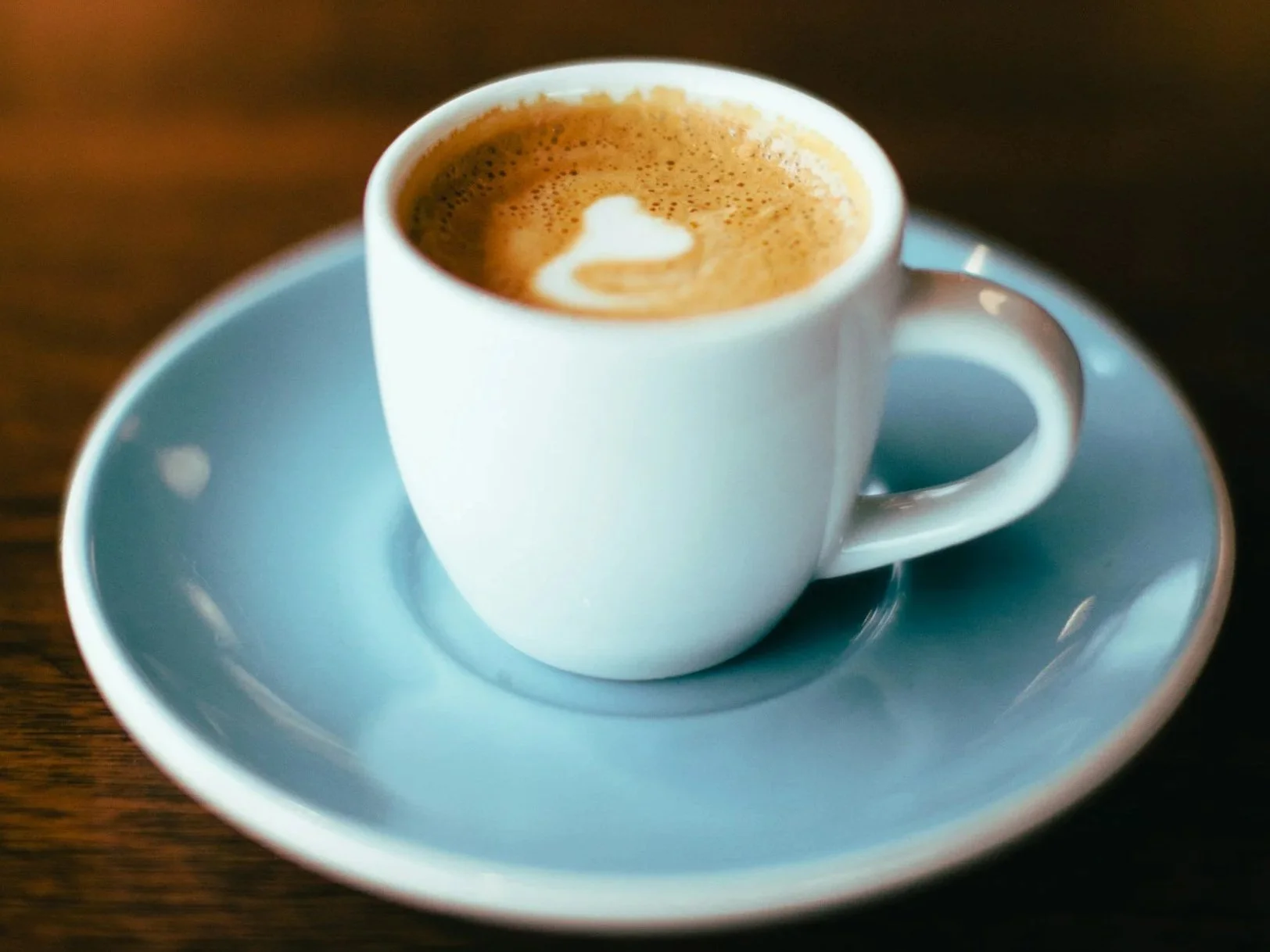 A blue ceramic cup of coffee with foam art on top, placed on a matching white saucer on a wooden table.
