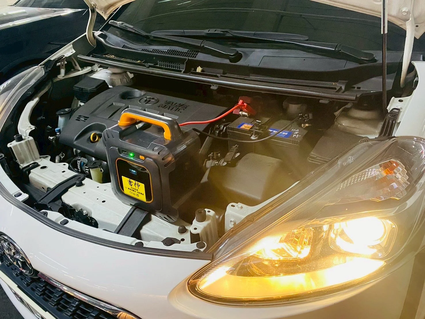 Open car hood showing engine bay with a portable battery charger connected to the car's battery.