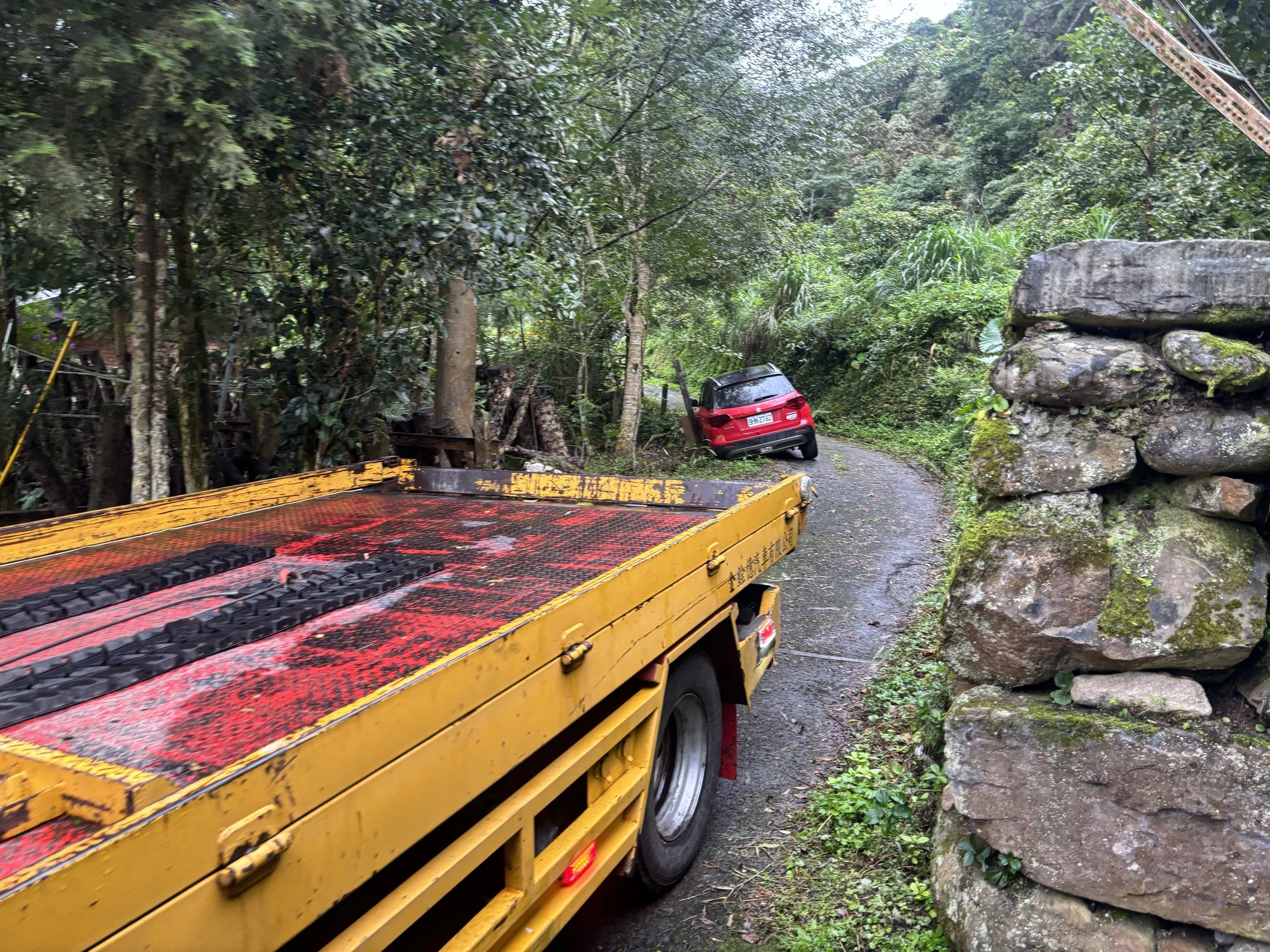 A narrow, winding road in a lush, green forest with a yellow tow truck on the left and a red car partially off the road in the background. Large rocks are on the right side of the road.