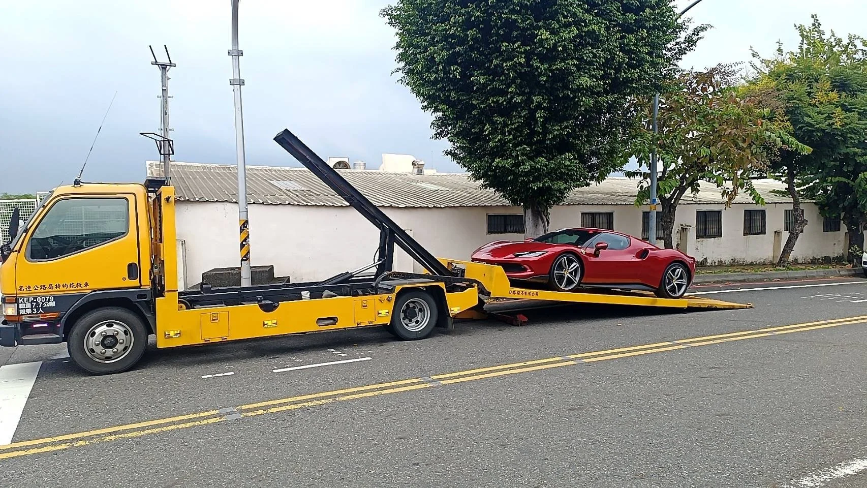 A red sports car on a yellow flatbed tow truck parked on a street with trees and a white building in the background.