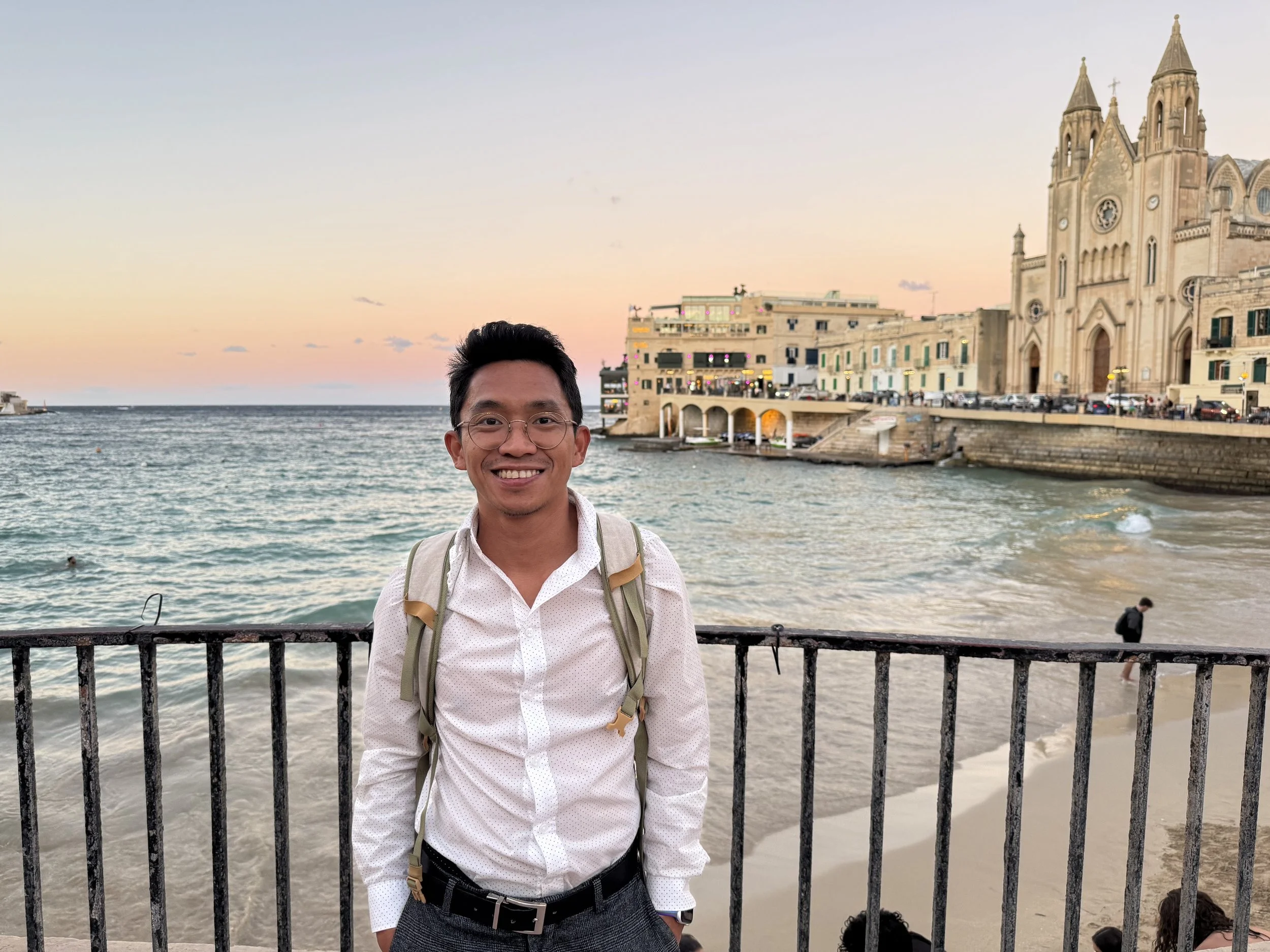 A smiling man with glasses and a backpack standing in front of a seaside view at sunset, with a church and buildings in the background.