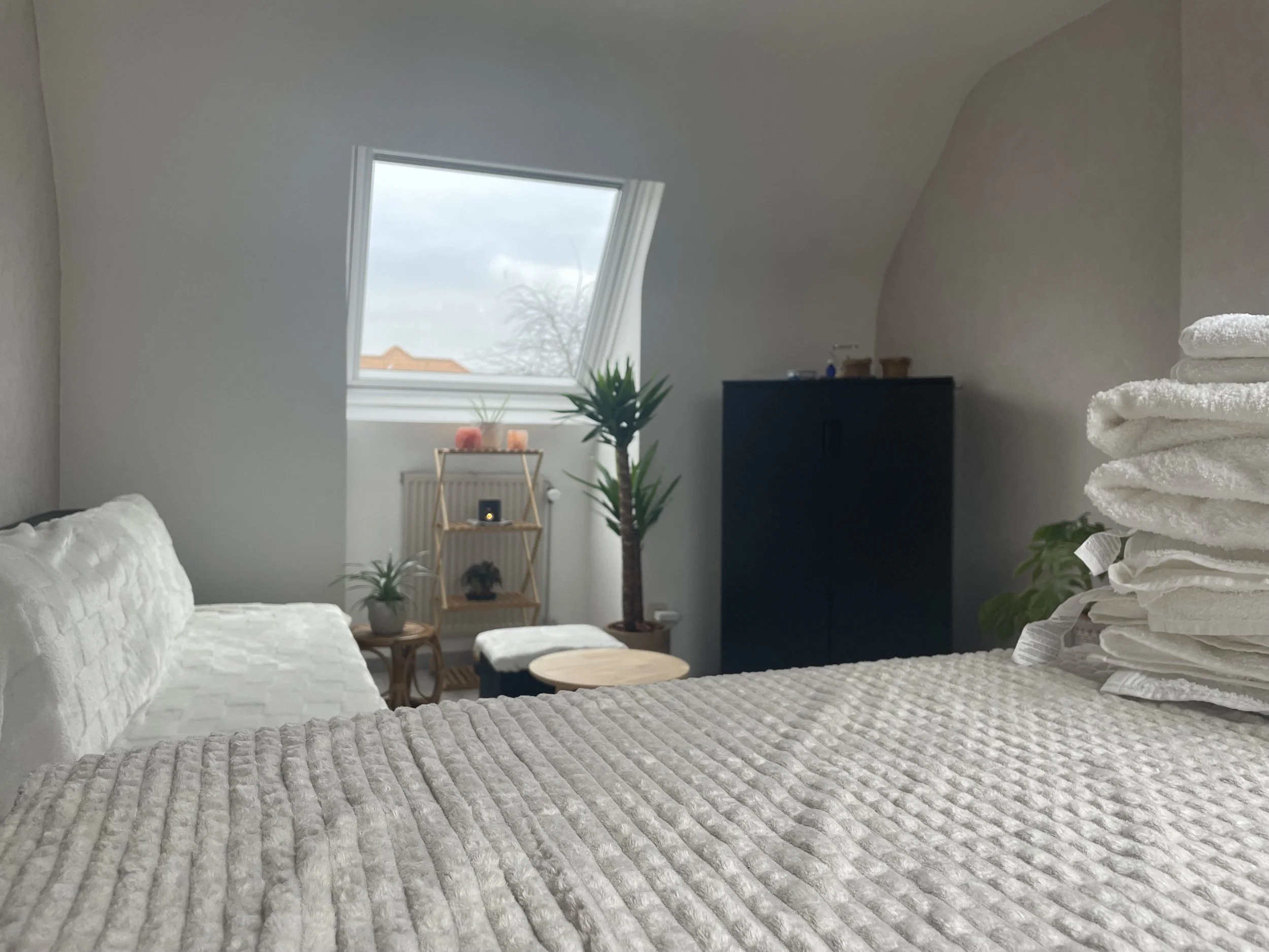 Cozy bedroom with a textured blanket on the bed, a stack of folded towels on the right, and a window with a view of the cloudy sky outside. Green plants and decorative items are placed around the room.