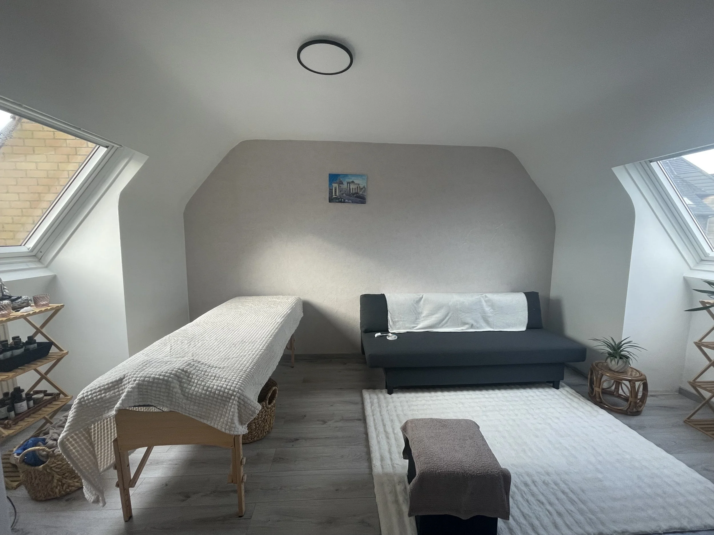 A therapy or massage room in an attic space with sloped ceiling and two large skylight windows. The room has a massage table on the left, a small black couch on the right with a white towel draped over the back, a small stool or ottoman in front, a white textured rug, a small wooden side table with a plant, and a shelving unit with skincare products. A small painting hangs on the wall above the couch, and a round ceiling light is installed on the ceiling.