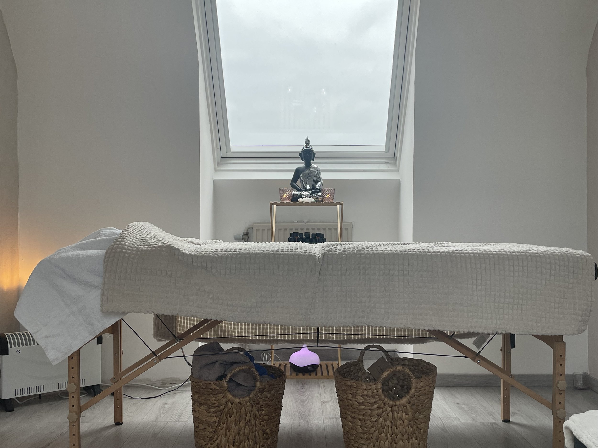 A massage table covered with a white towel in a room with a large skylight window. There are two wicker baskets beneath the table, containing towels or cloths. On the window sill, there is a black Buddha statue and a small table with candles or decorative items. A purple light diffuser is also visible beneath the table.