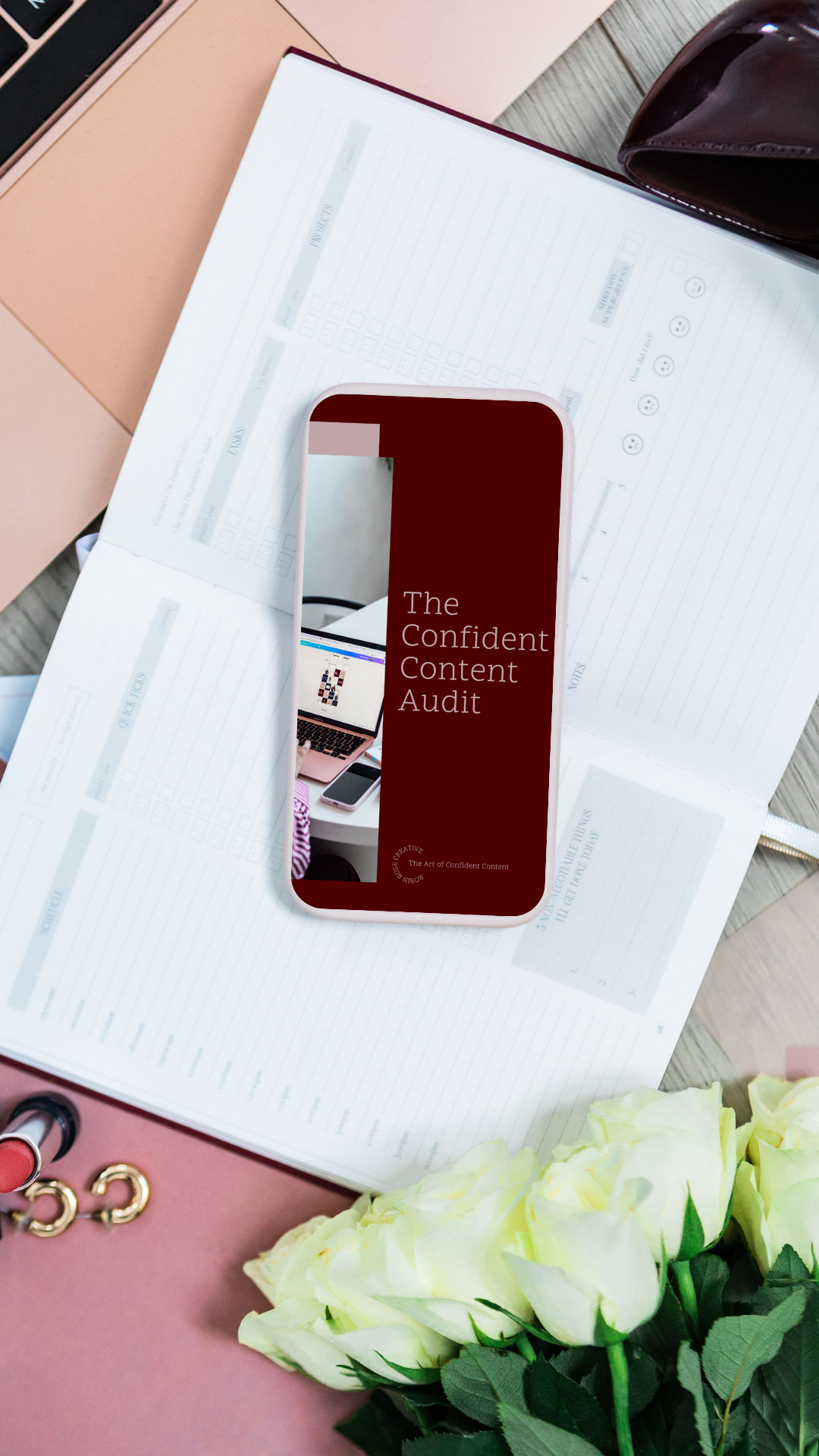 A phone displaying the text 'The Confident Content Audit' resting on a desk with papers, a black wallet, and a partially visible laptop. A bouquet of white roses is in the foreground.