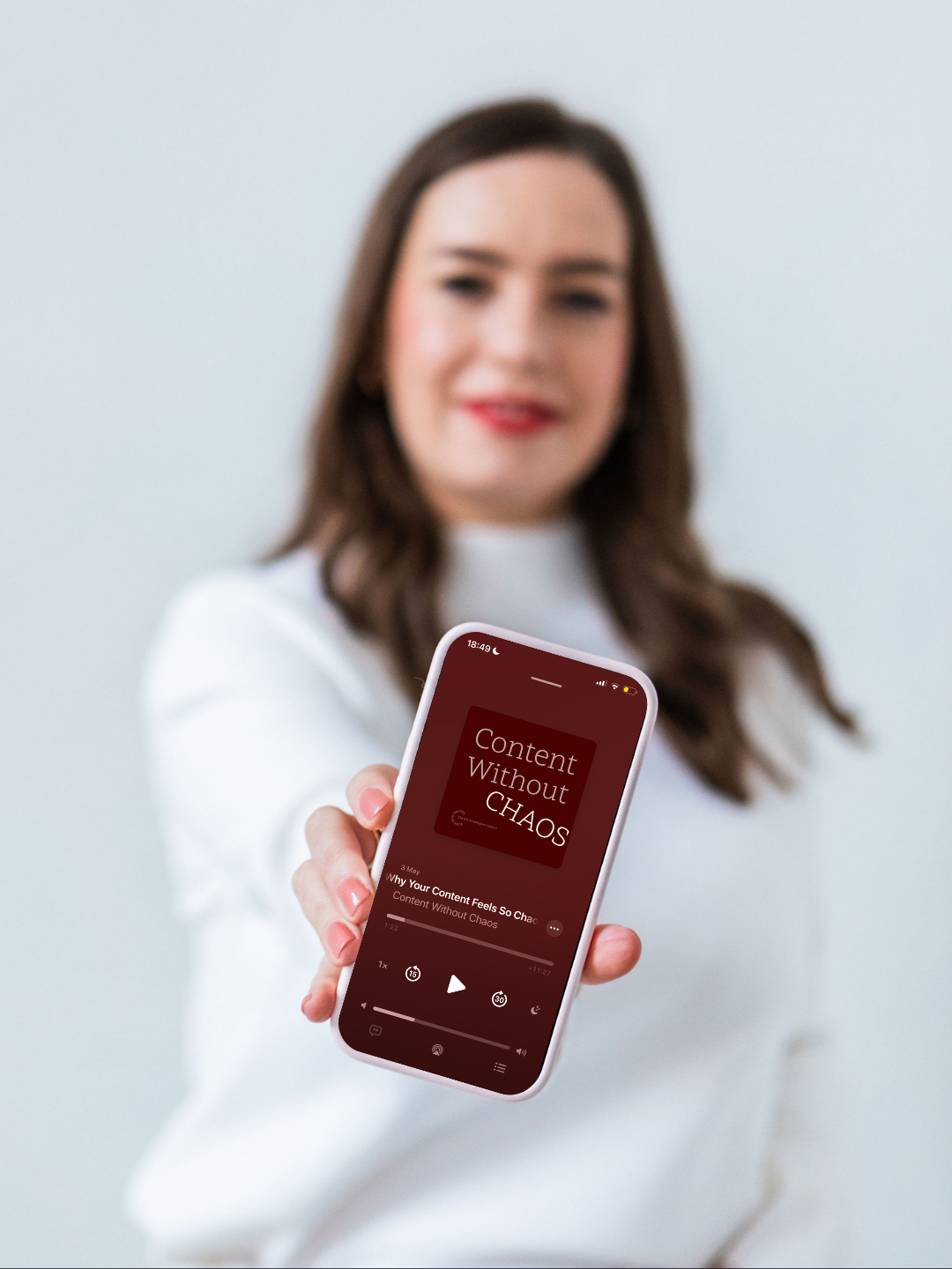 A woman in white shirt smiling and holding a smartphone showing a podcast titled 'Content Without Chaos.'