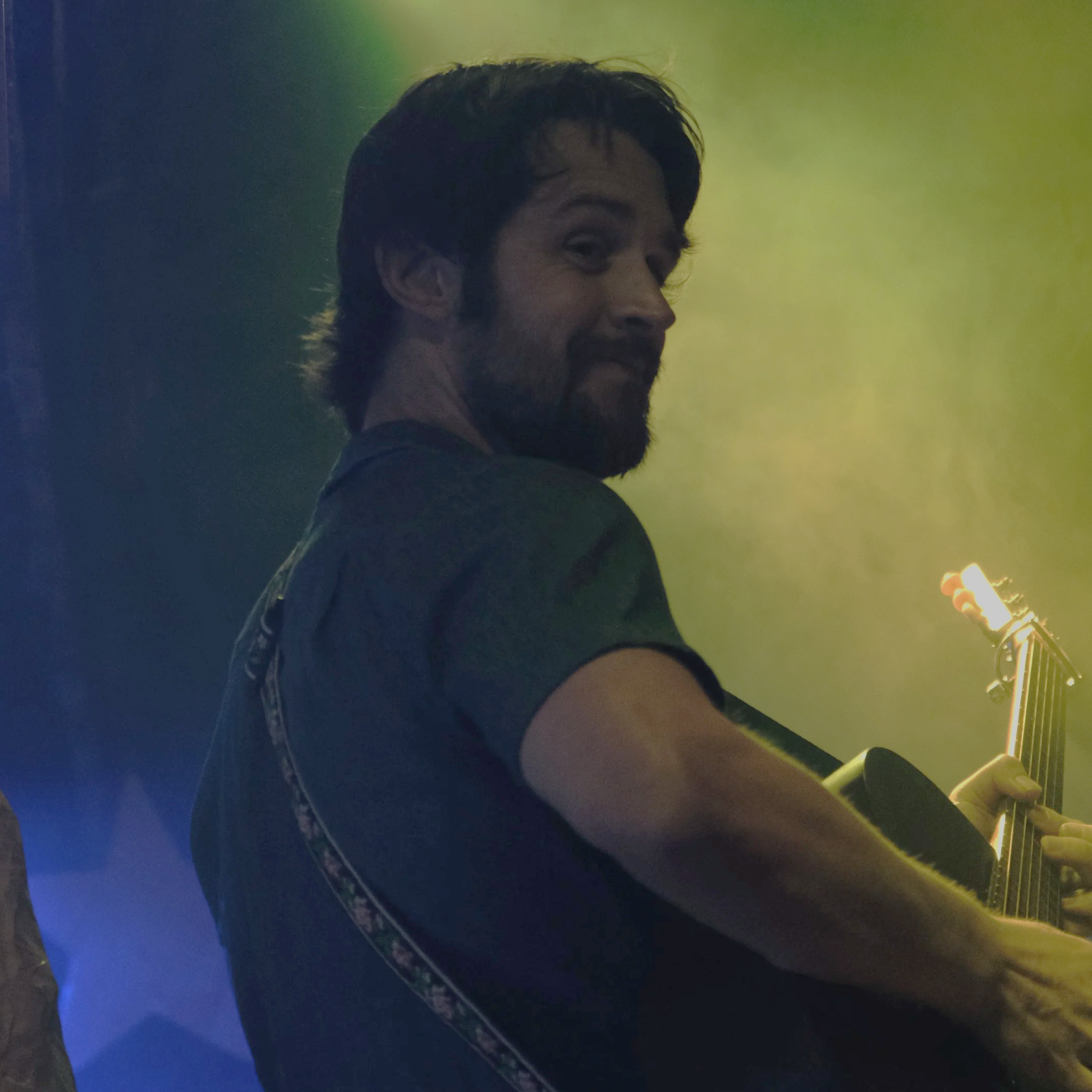 A man with dark hair and a beard is playing guitar on stage, smiling and looking at the camera, with colorful stage lights behind him.