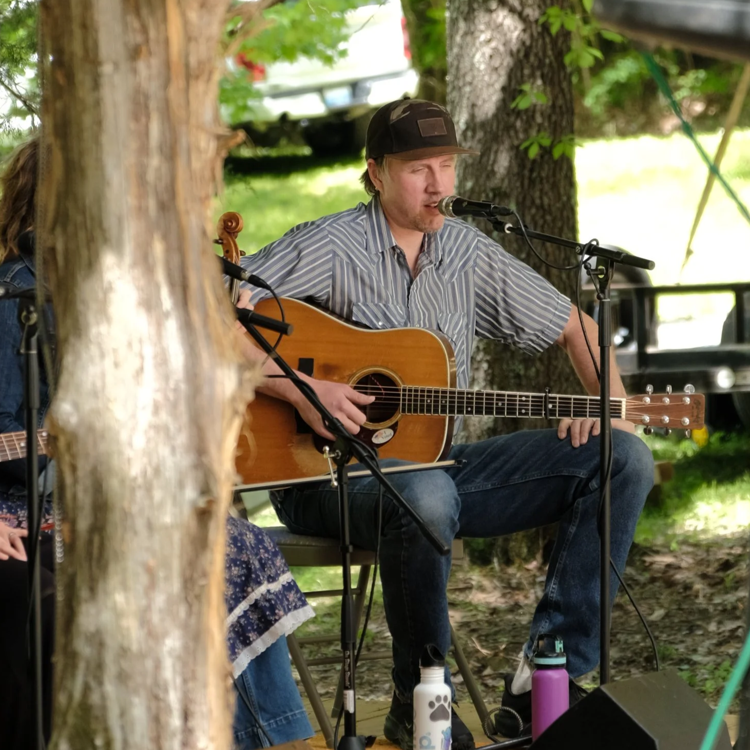A woman with shoulder-length black hair smiling outdoors with green plants in the background.