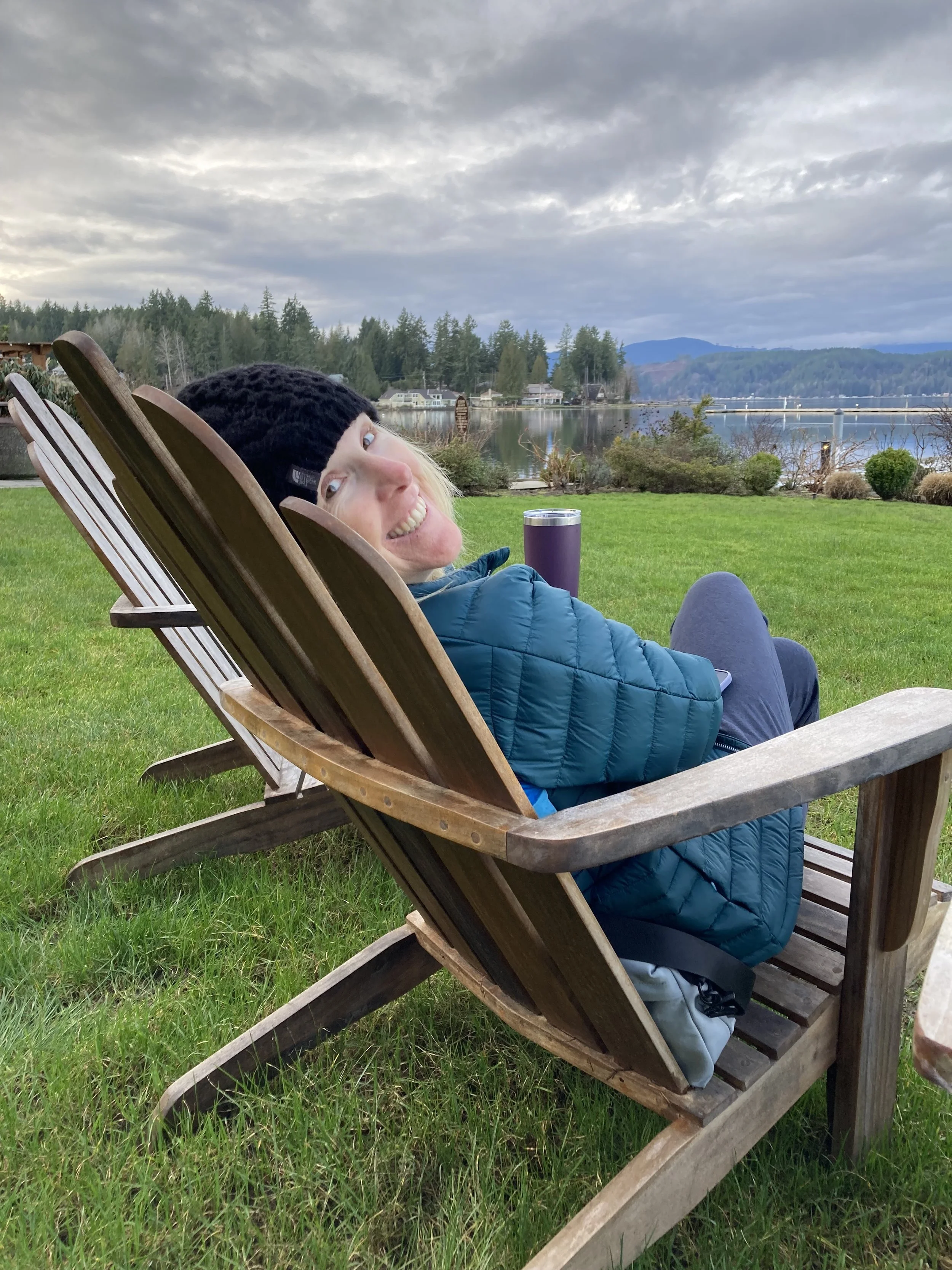 Woman sitting on wooden lawn chair near a lake, smiling, wearing a black knit hat and blue jacket, with a purple tumbler nearby, scenic background of trees and mountains under cloudy sky.