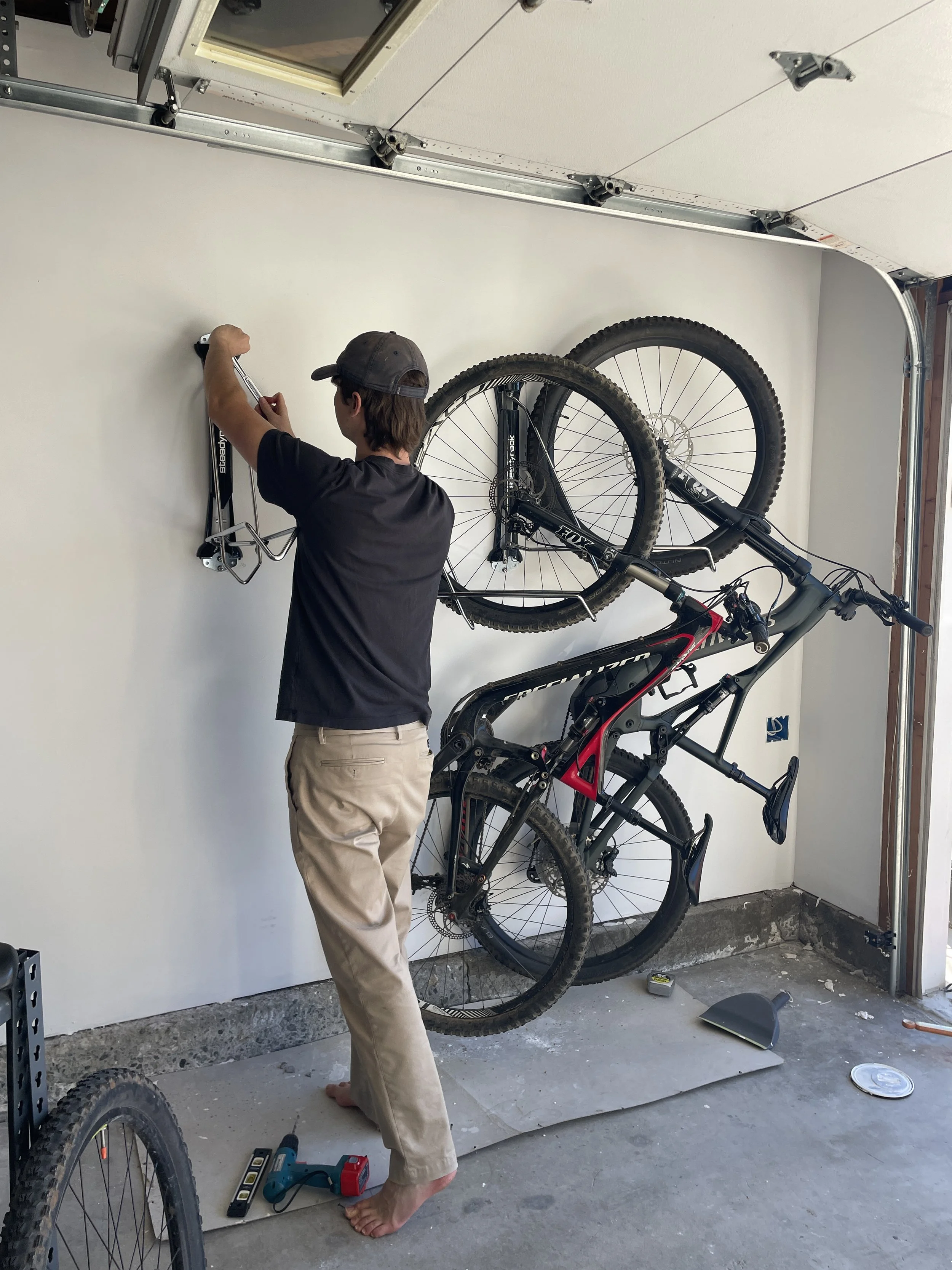 A person standing barefoot in a garage, installing or adjusting a bike wall mount for two mountain bikes, with tools and bike accessories on the floor.