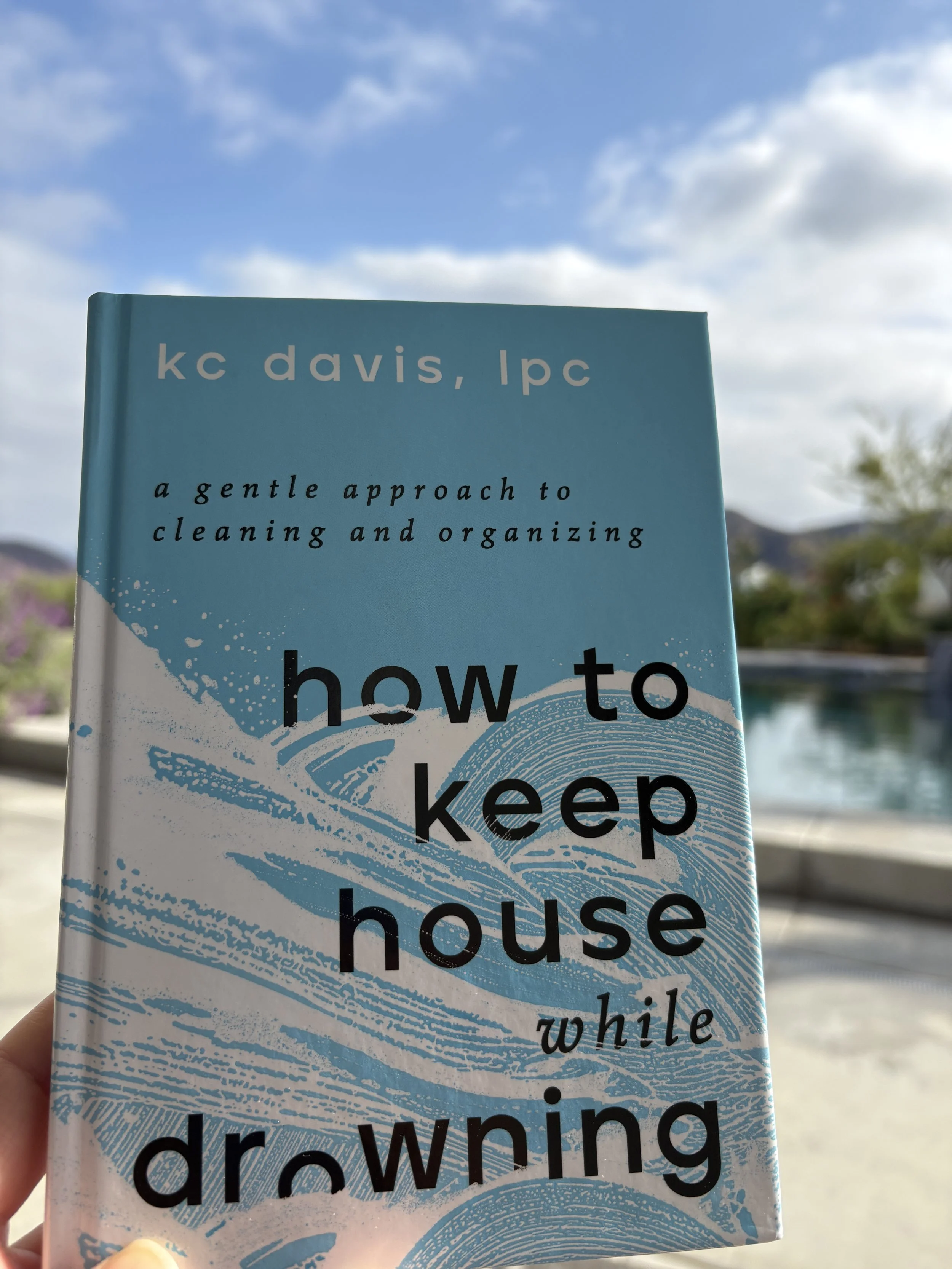 A book titled 'How to Keep House While Drowning' by KC Davis, the cover features a blue and white design with water waves, held outdoors with a blurry background of a pool and sky with clouds.