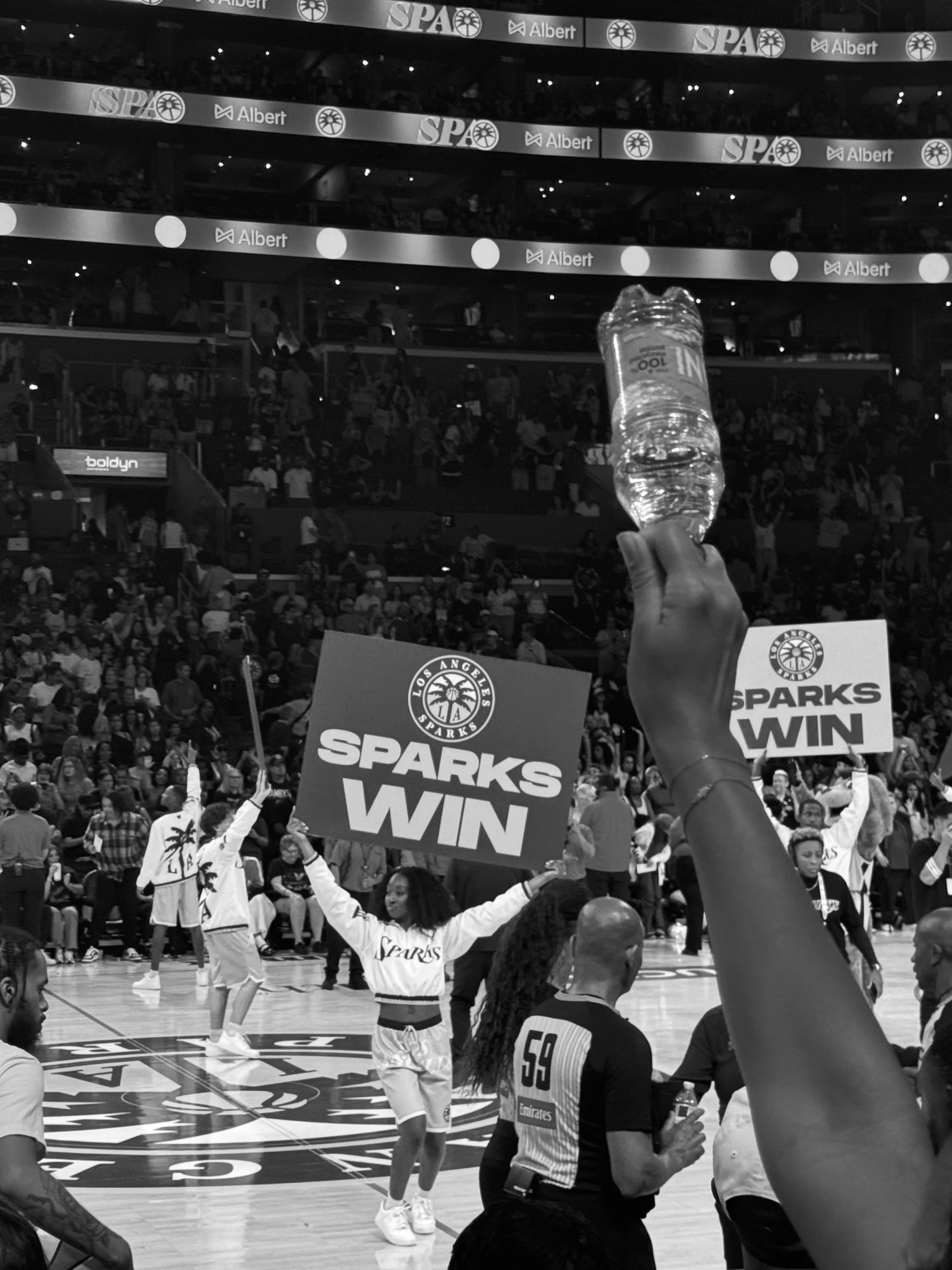 A basketball game celebration scene on the court of the Los Angeles Sparks team, with a woman holding a sign reading 'SPARKS WIN' and a large crowd in the stands.