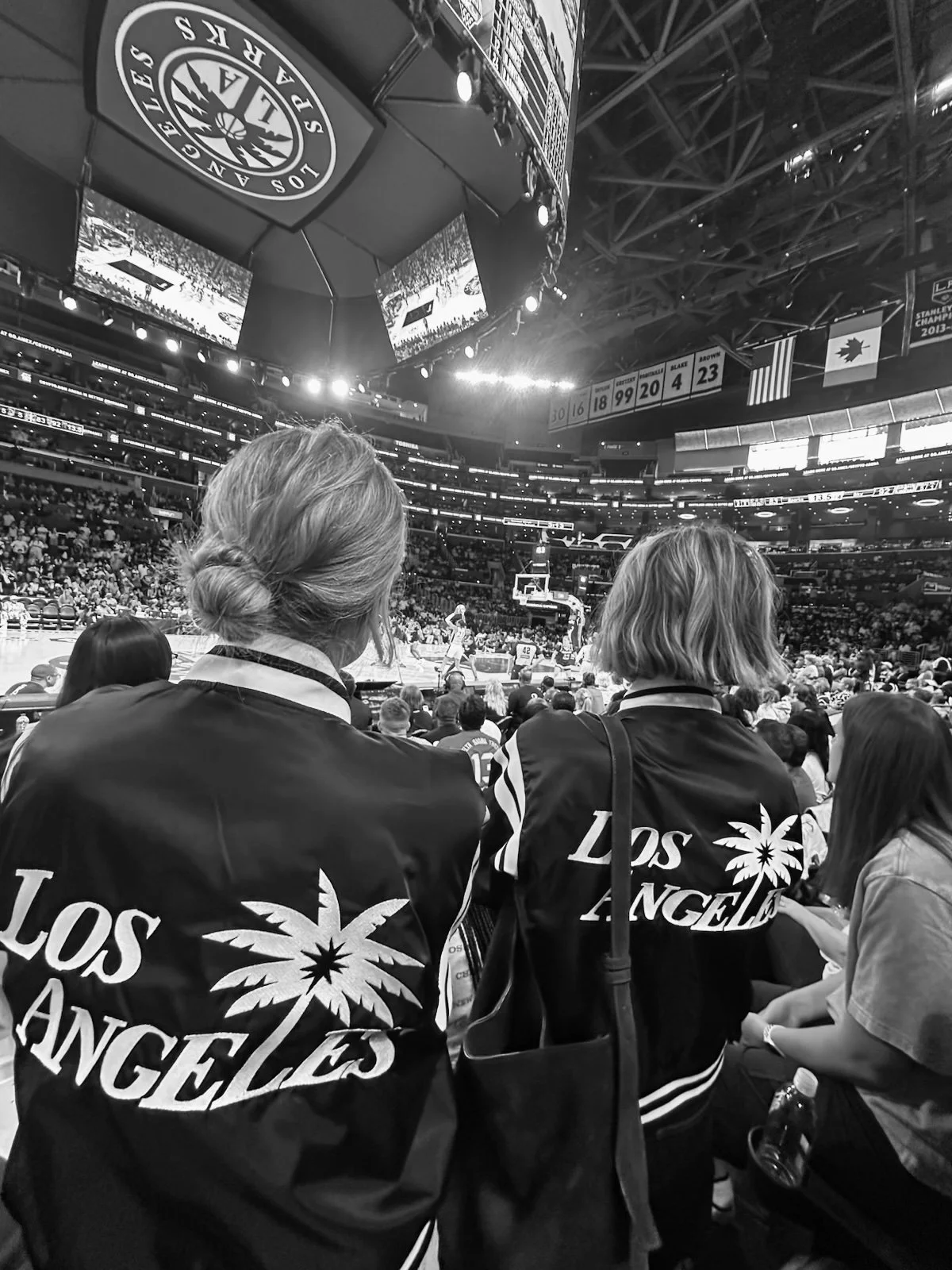 Two women wearing Los Angeles jackets watch a basketball game in an arena filled with spectators.
