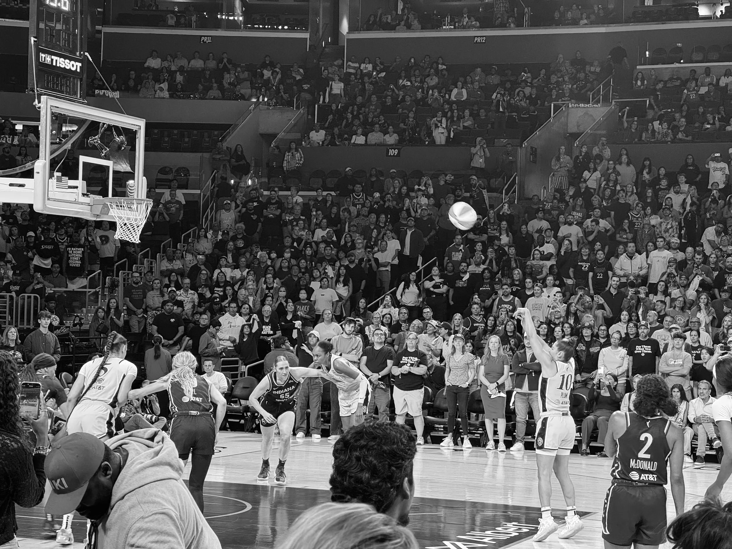 A women's basketball game in progress on a court with players in action, with one player shooting the ball and others defending, surrounded by spectators in the stands.