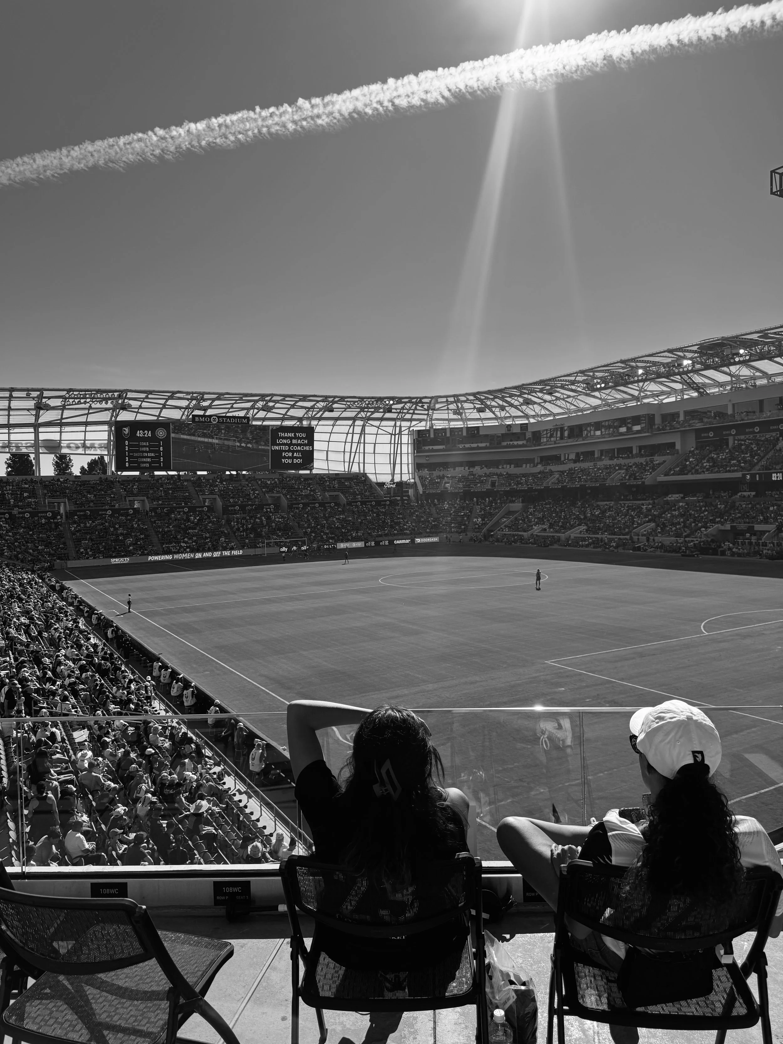 Two women sitting in front of a soccer stadium, watching a game on the field, with a crowd in the stands and a clear sky overhead.