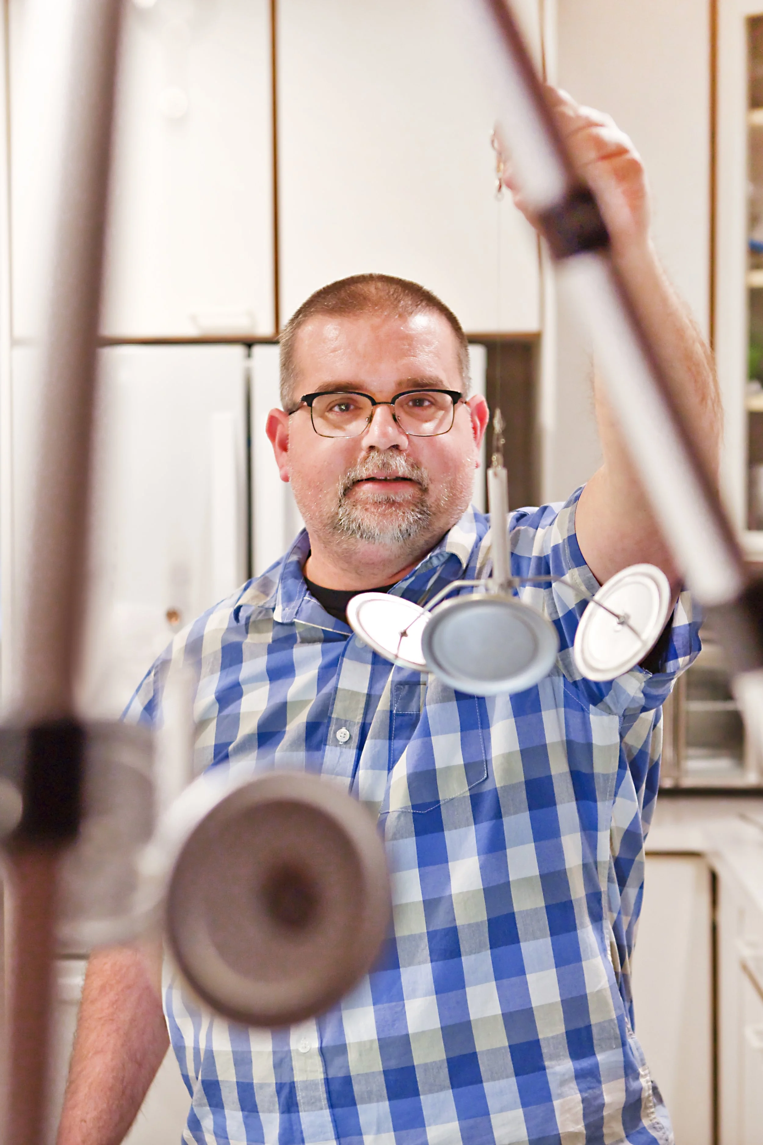 A man with glasses and a checkered shirt is inspecting pipes or tubes in a workshop or kitchen.