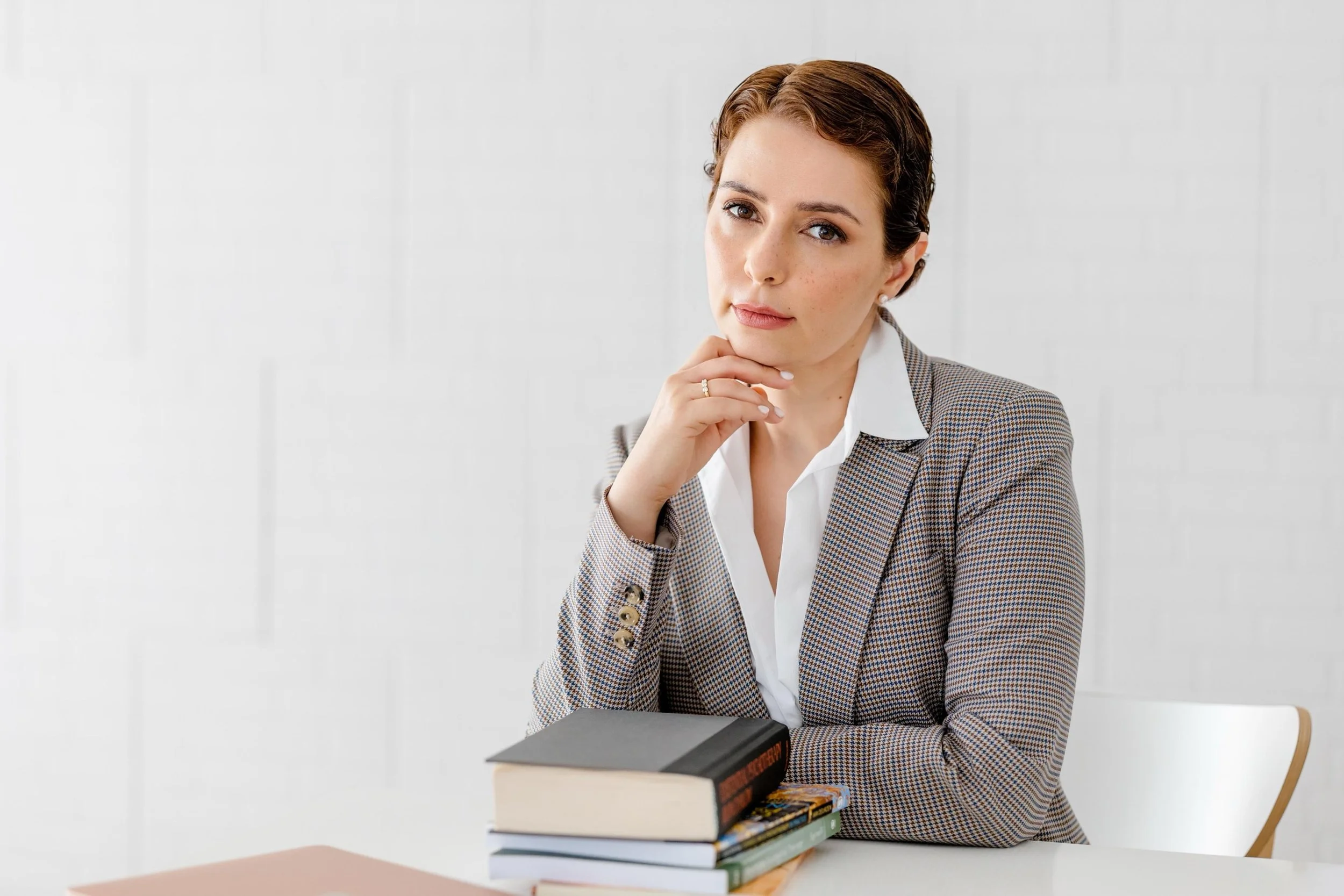 Mulher jovem com cabelo castanho, vestida com blazer xadrez e camisa branca, sentada à mesa com livros empilhados à sua frente, olhando com expressão pensativa.