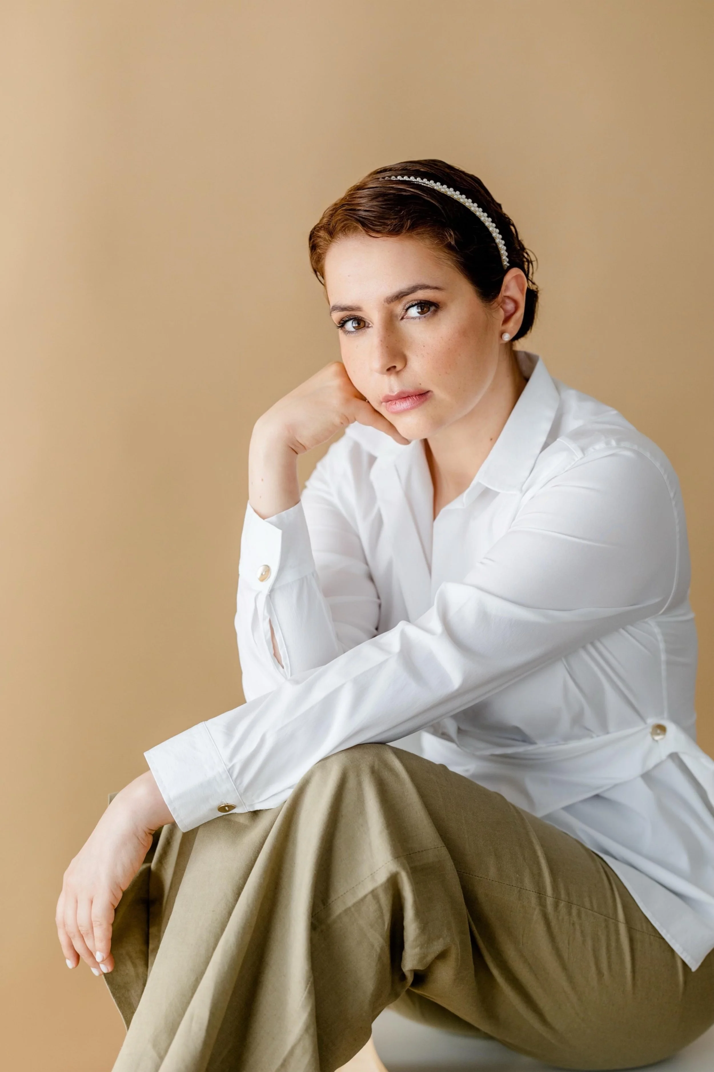 Mulher com cabelo castanho, usando tiara de pérolas, camiseta branca e calças cáqui, sentada com o queixo na mão, olhando diretamente para a câmera, contra fundo bege.