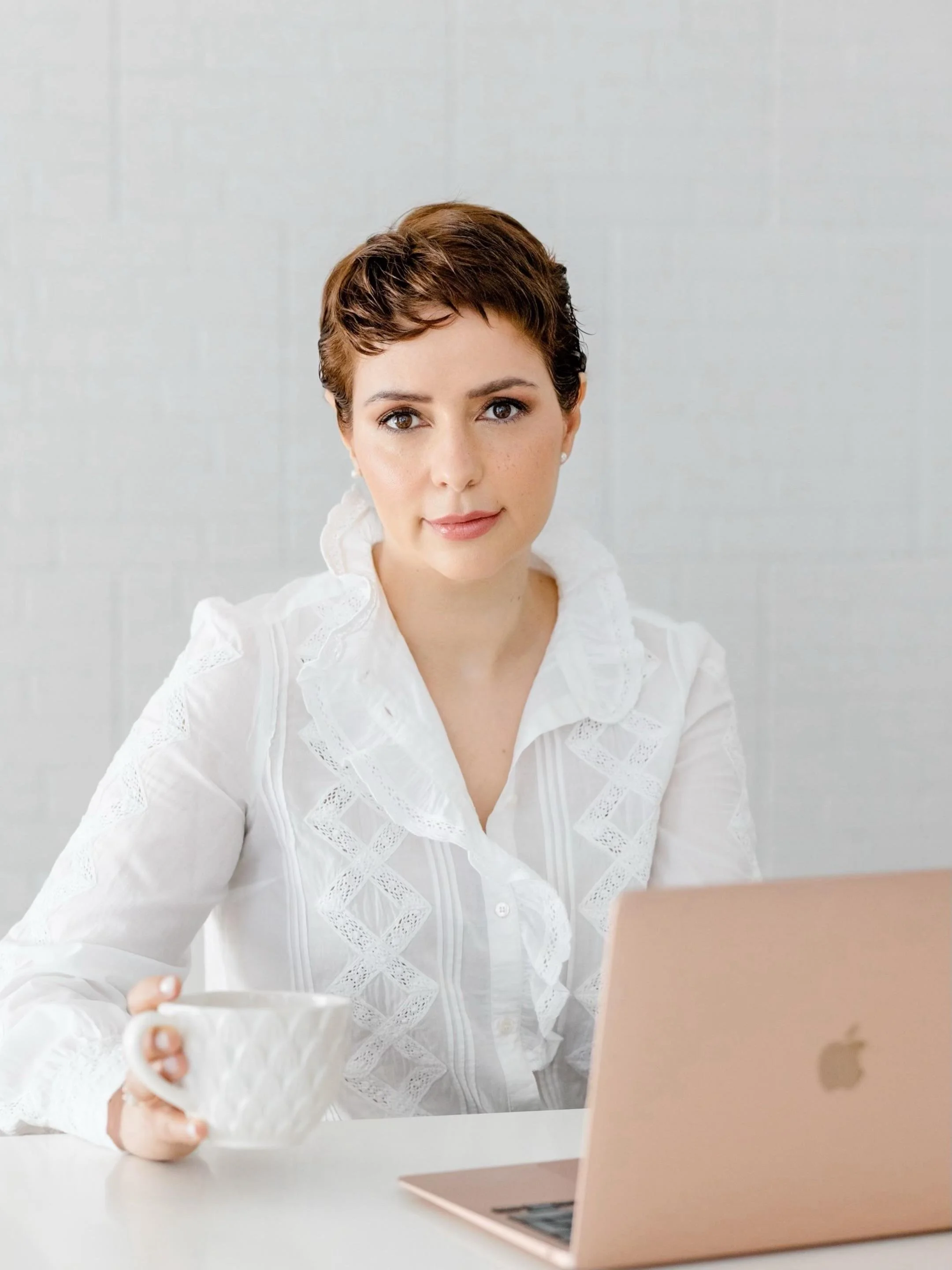 Mulher com cabelo curto e castanho, vestindo blusa branca com detalhes em renda, segurando uma xícara branca, sentada em frente a um computador rosé, em um ambiente bem iluminado.