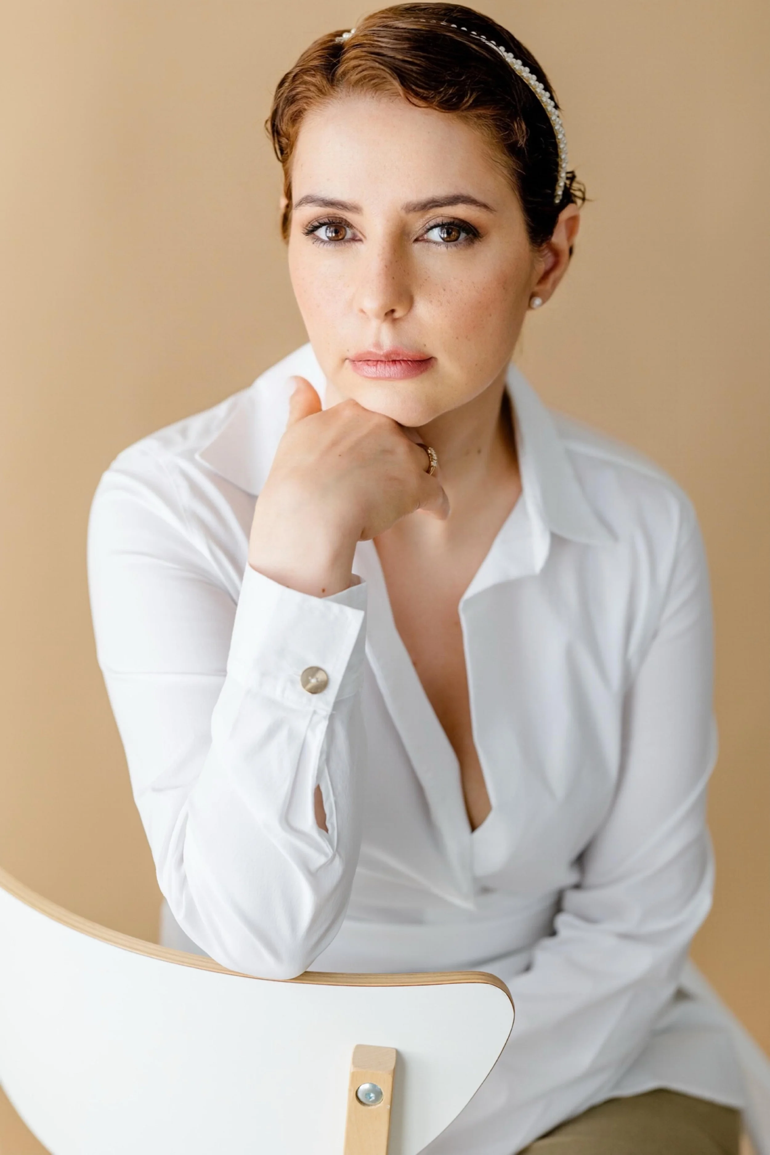 Mulher com cabelos castanhos curtos, usando blusa branca e acessórios de cabeça, sentada em uma cadeira branca com fundo bege