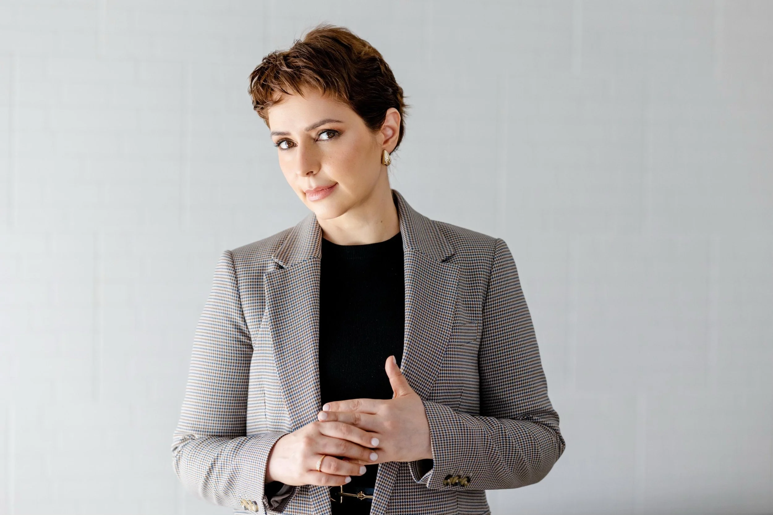 Mulher com cabelo curto e castanho, usando blazer de xadrez cinza com preto e blusa preta, posando com as mãos juntas diante do peito, contra parede cinza lisa.