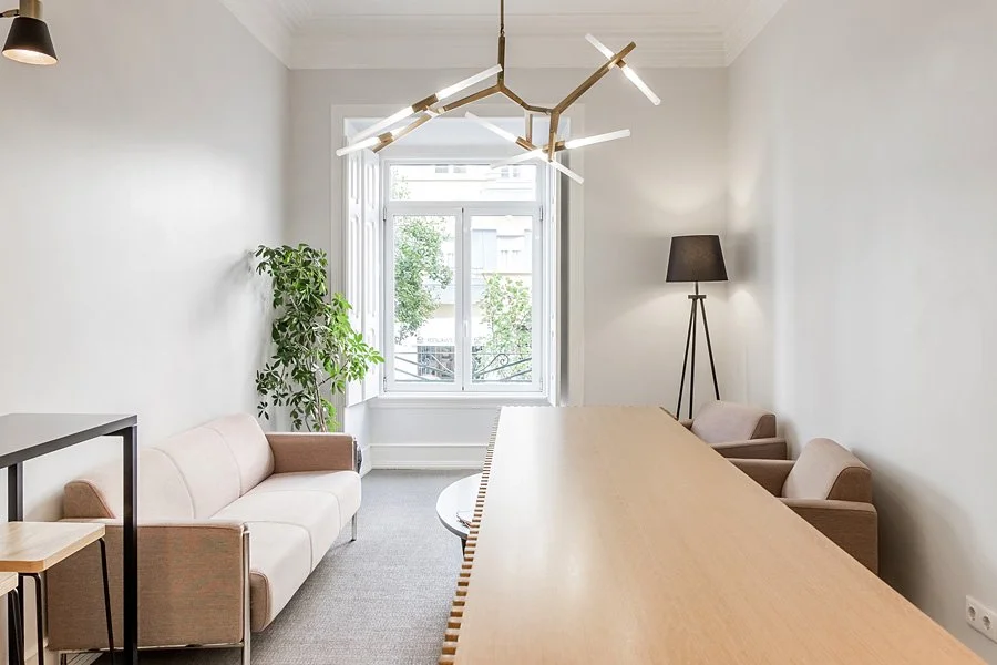 Sala de reunião com mesa de madeira, poltronas, sofá bege, luminária de chão, luminária moderna no teto, janela grande com vista para árvores.