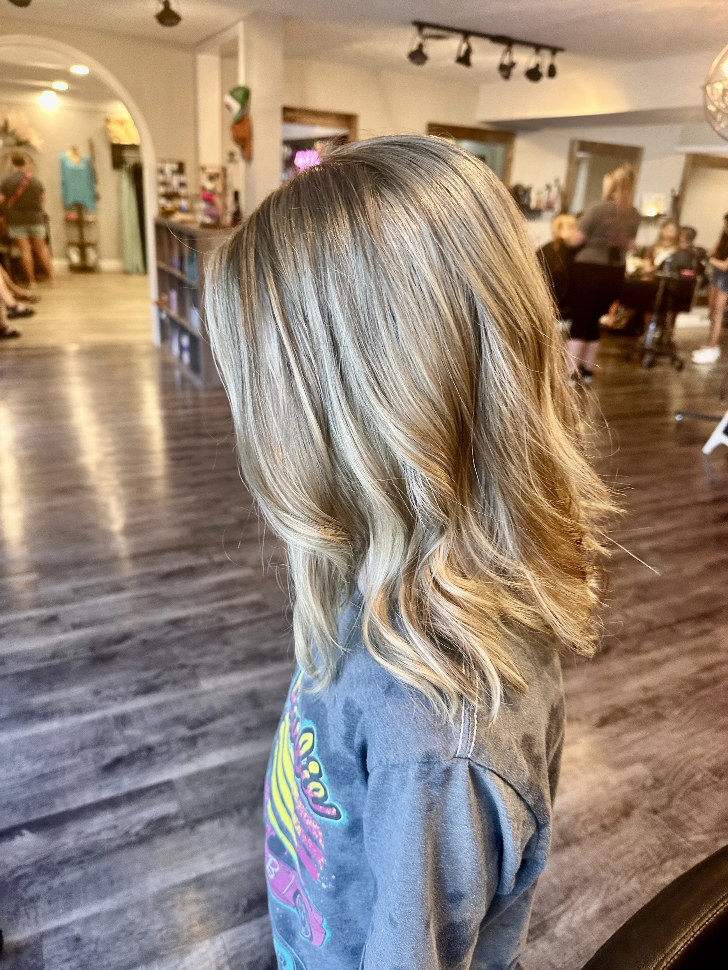 Side view of a young girl with blonde, wavy hair, wearing a gray T-shirt with colorful text and graphics, in a hair salon or similar setting.