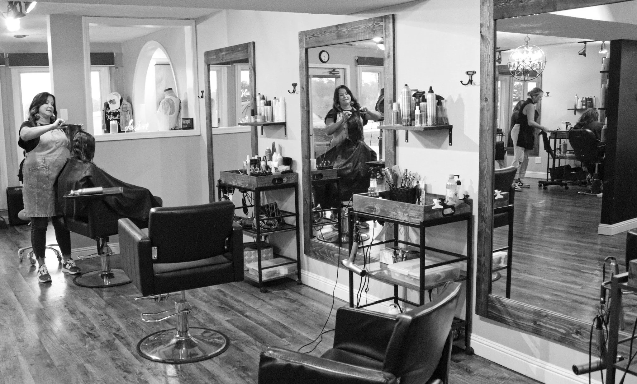 Inside a hair salon with two hairstylists cutting clients' hair, large mirrors on the wall, and salon chairs on a wooden floor.
