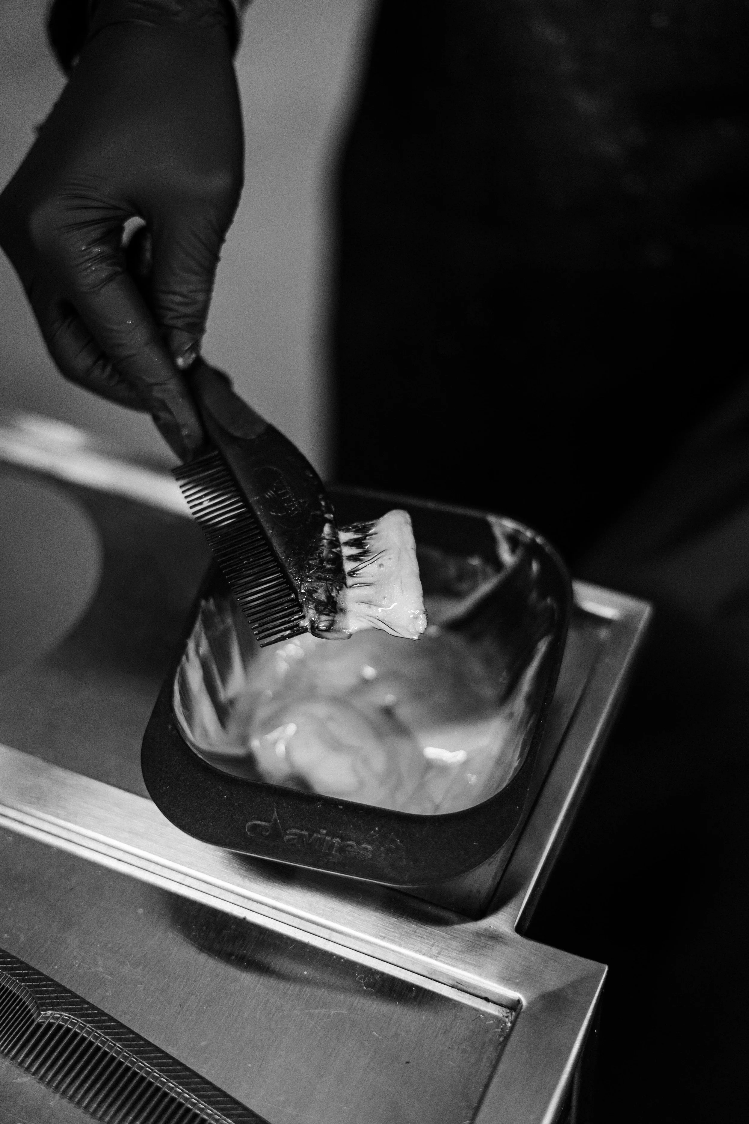 A person wearing black gloves combs a piece of hair in a black silicone container on a stainless steel ashtray.