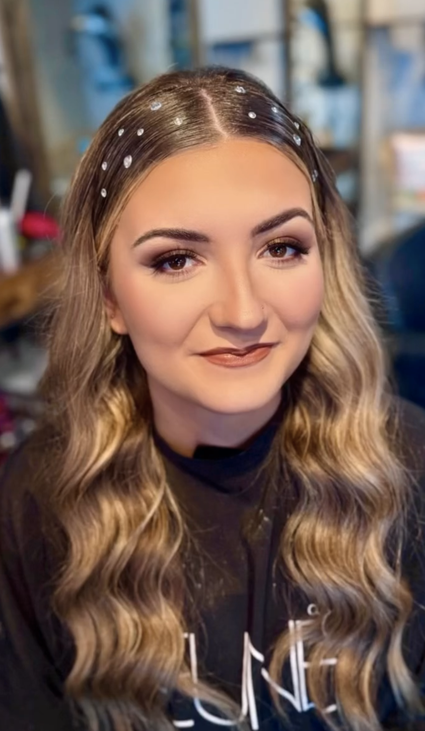 A young woman with long, wavy blonde hair adorned with small pearls, wearing makeup and a dark top, smiling at the camera in an indoor setting.