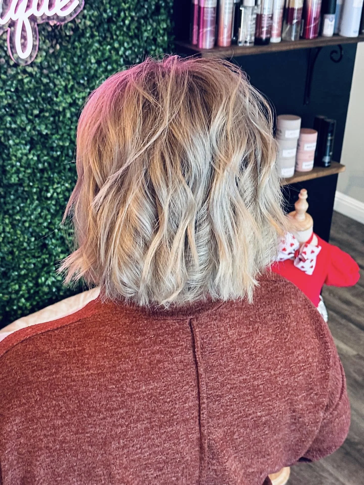 Back view of a woman with wavy blonde hair sitting in a salon chair, with a green wall and shelves of hair products in the background.