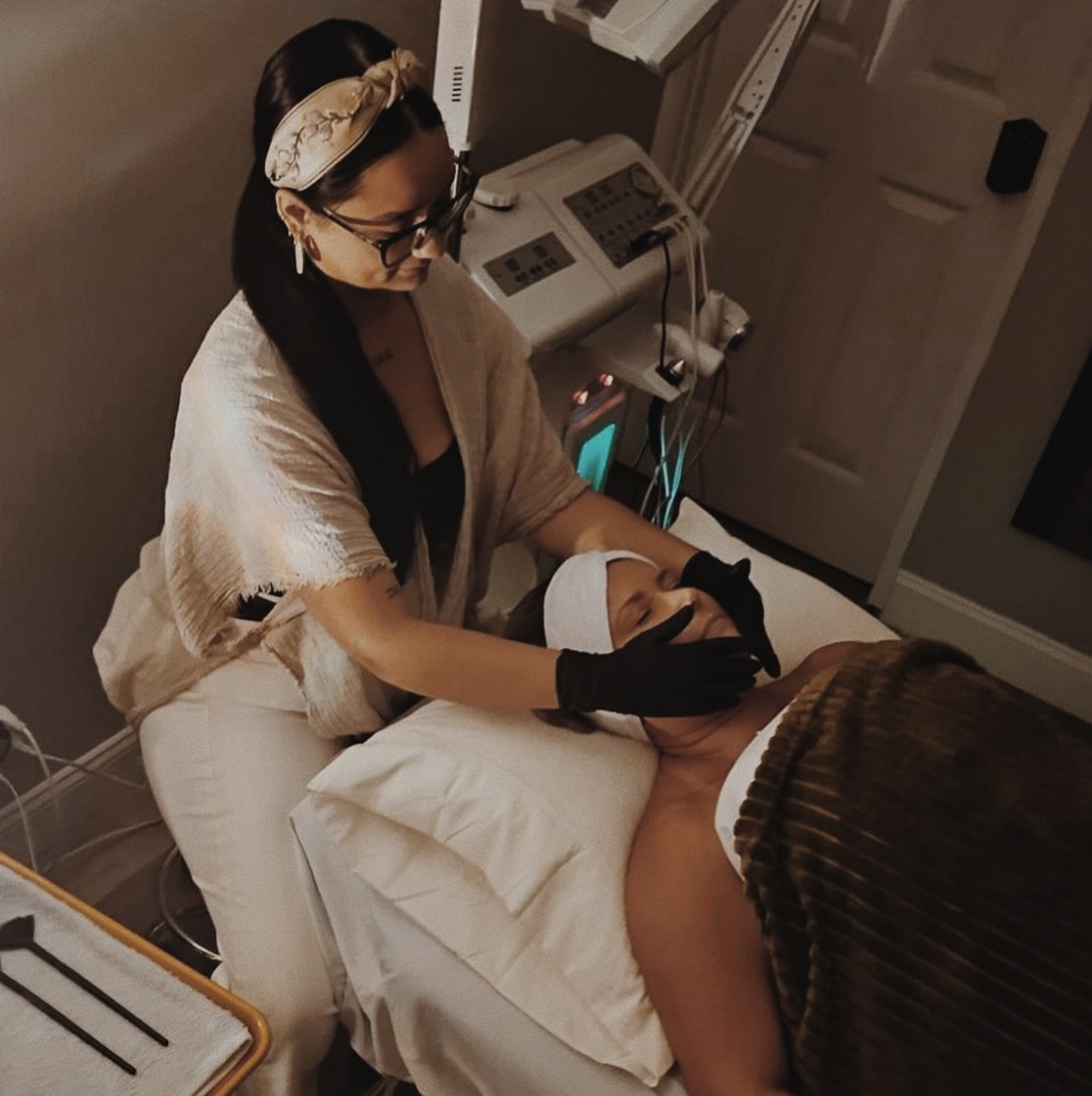 A woman with glasses, wearing a beige headband and black gloves, is performing a facial treatment on a woman lying on a bed, in a dimly lit room with medical equipment nearby.
