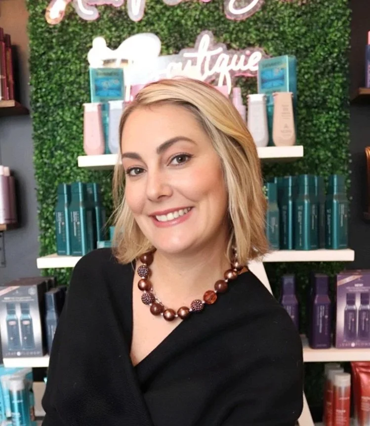 A woman with shoulder-length hair, smiling, wearing a black top and a beaded necklace, sitting in front of shelves with hair products and a neon sign that says 'Hair Flaw Salon' in a hair salon.