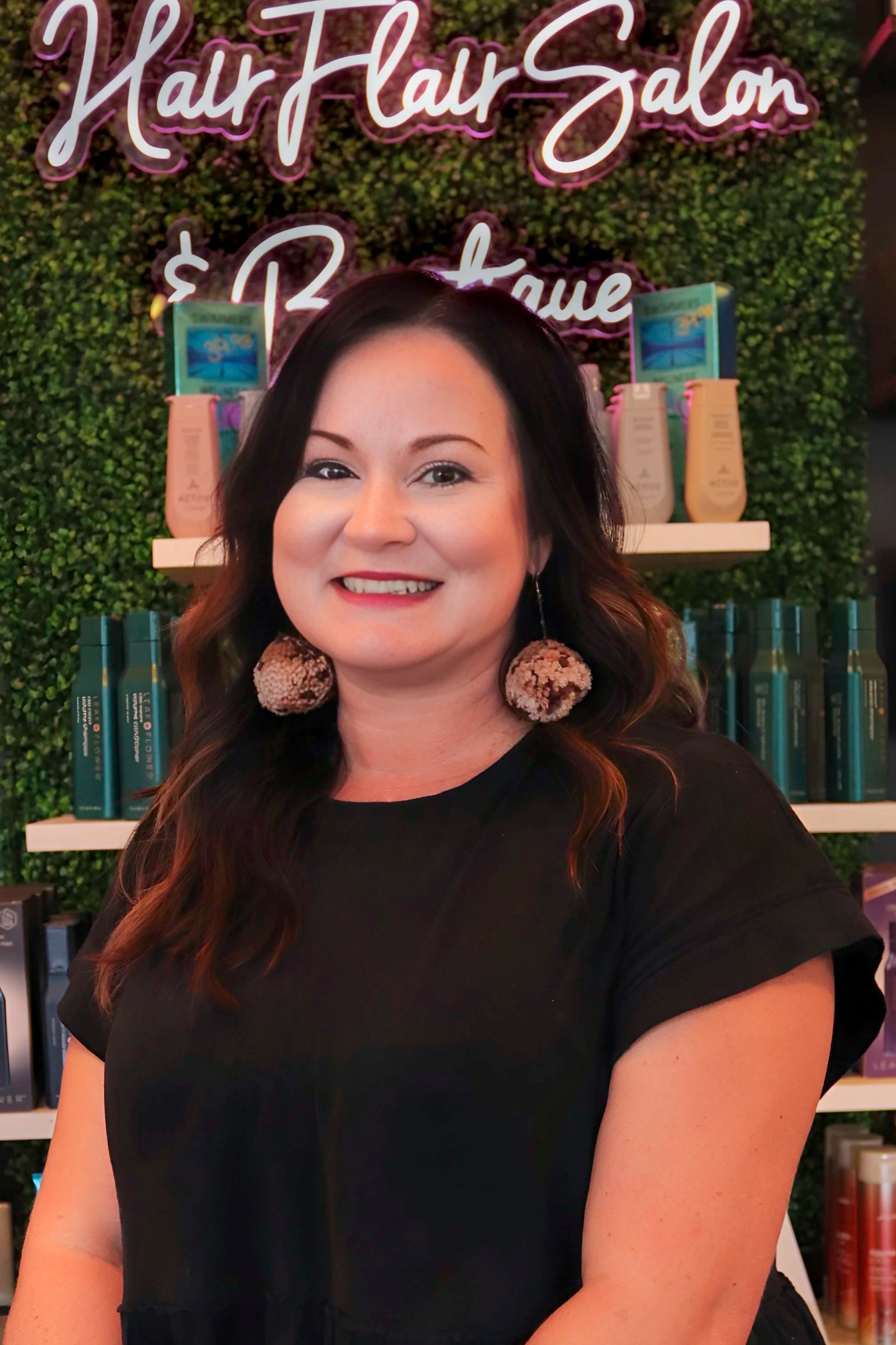 A smiling woman with wavy hair wearing large pom-pom earrings, standing in front of a hair salon display. The background includes products on shelves and a illuminated sign that reads 'Hair Hair Salon'.