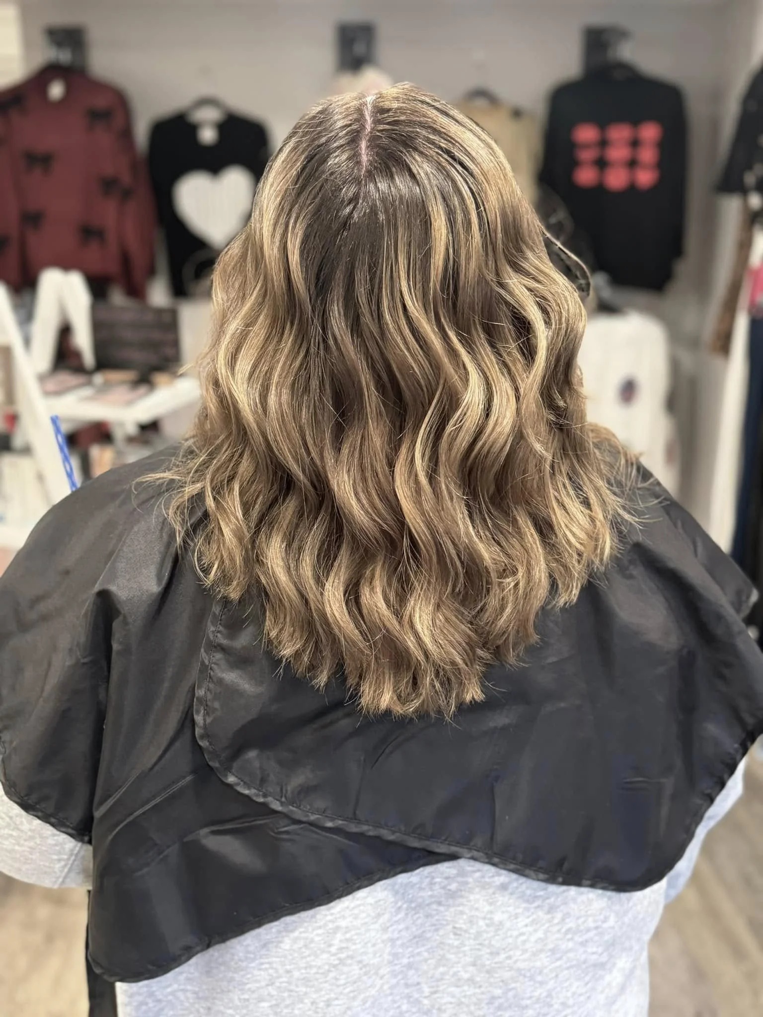 Back view of a woman with wavy, light brown hair with blonde highlights, sitting in a salon or boutique with clothing items hanging on the wall behind her.