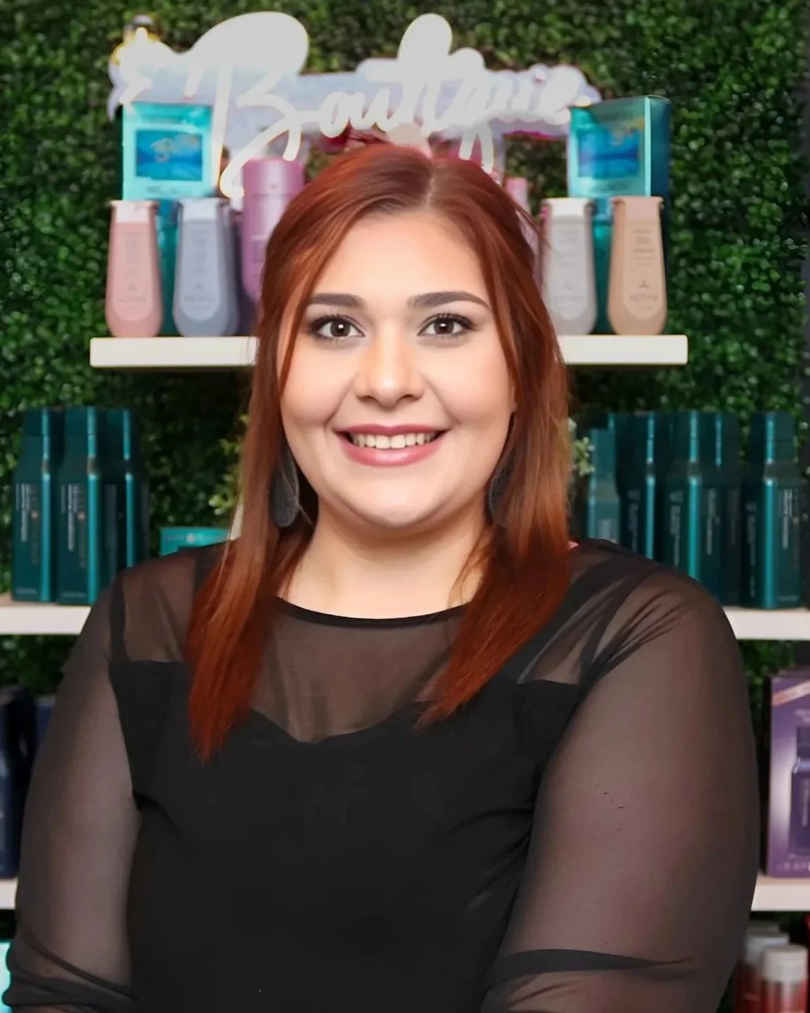 A smiling woman with shoulder-length hair in a retail store surrounded by shelves of hair care products and a decorative background.