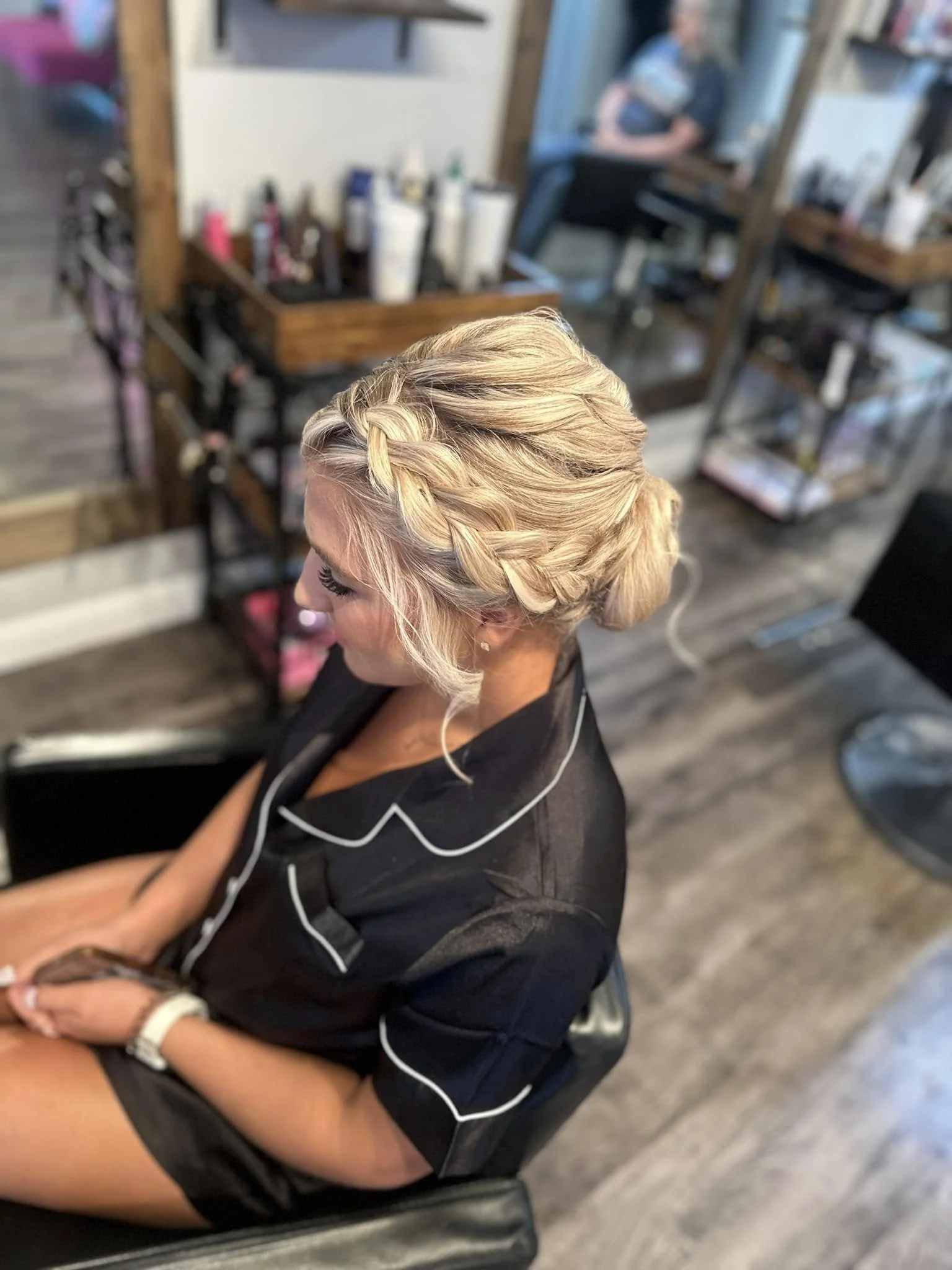 A woman with blonde hair styled in an elegant braided updo, sitting in a salon chair, wearing a black robe with white piping, in a hair salon with various hair products and tools visible in the background.