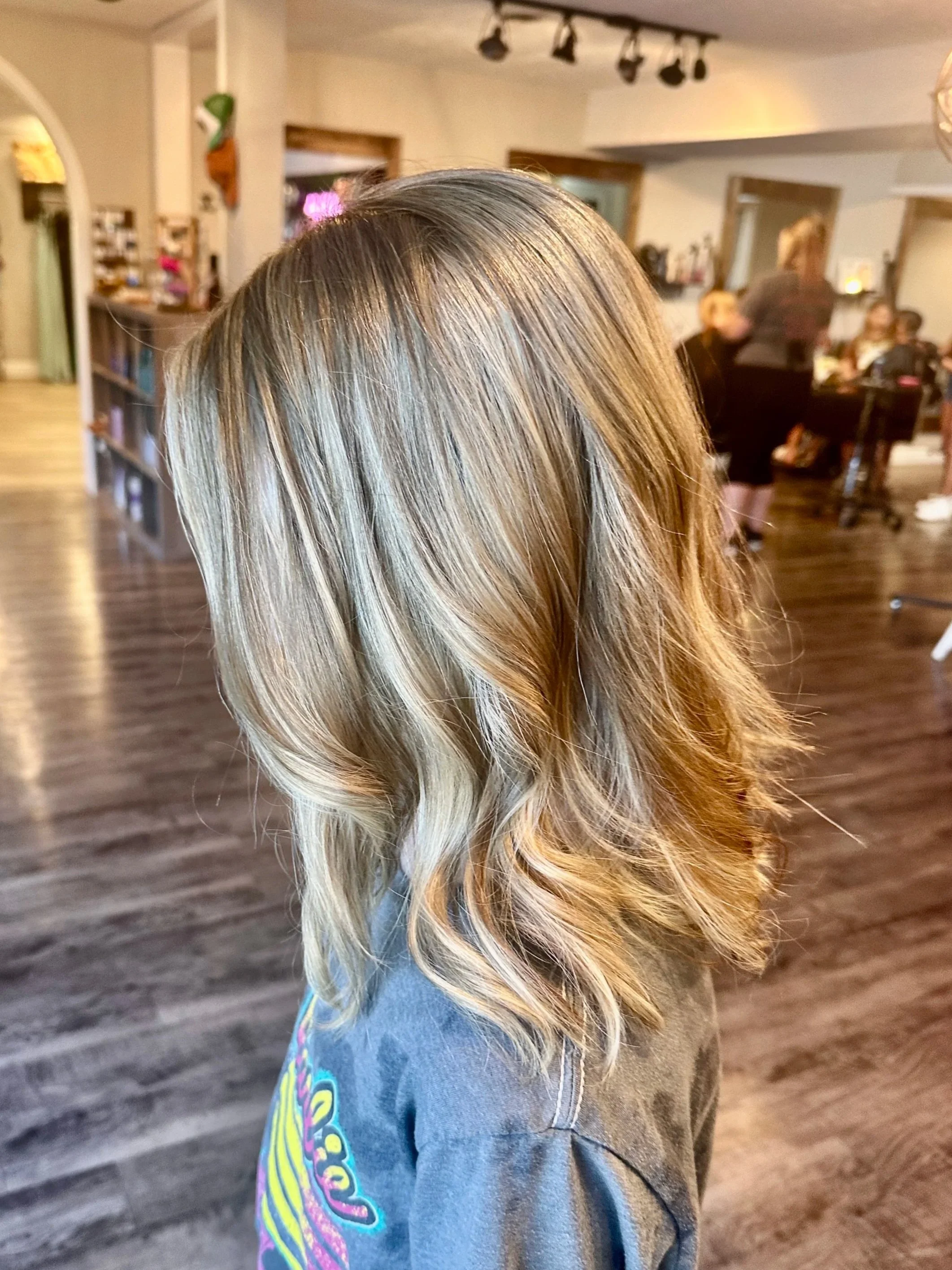 Side view of a woman with wavy blonde hair in a hair salon, with other clients and salon staff in the background.
