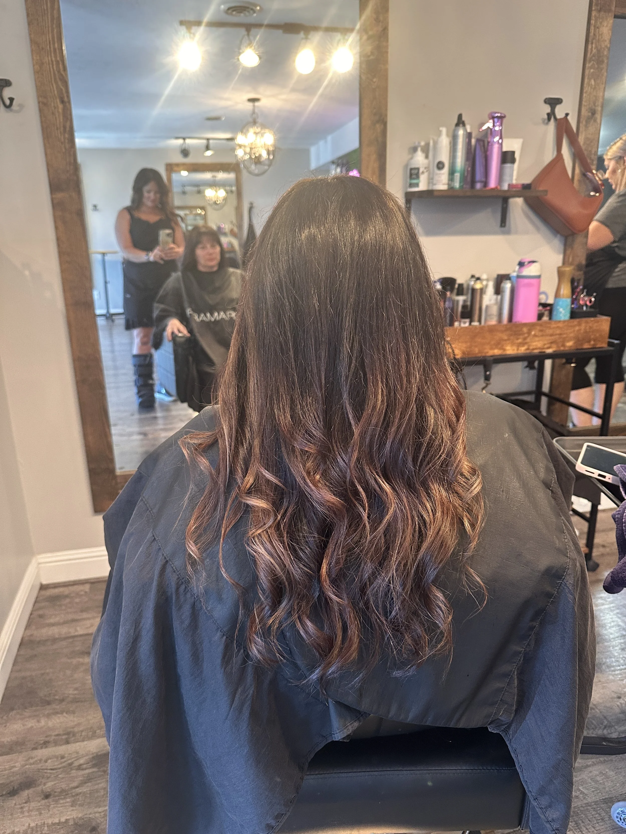 Back view of a woman with long, wavy, ombre hair sitting in a salon chair, with a mirror reflecting her and the salon interior.