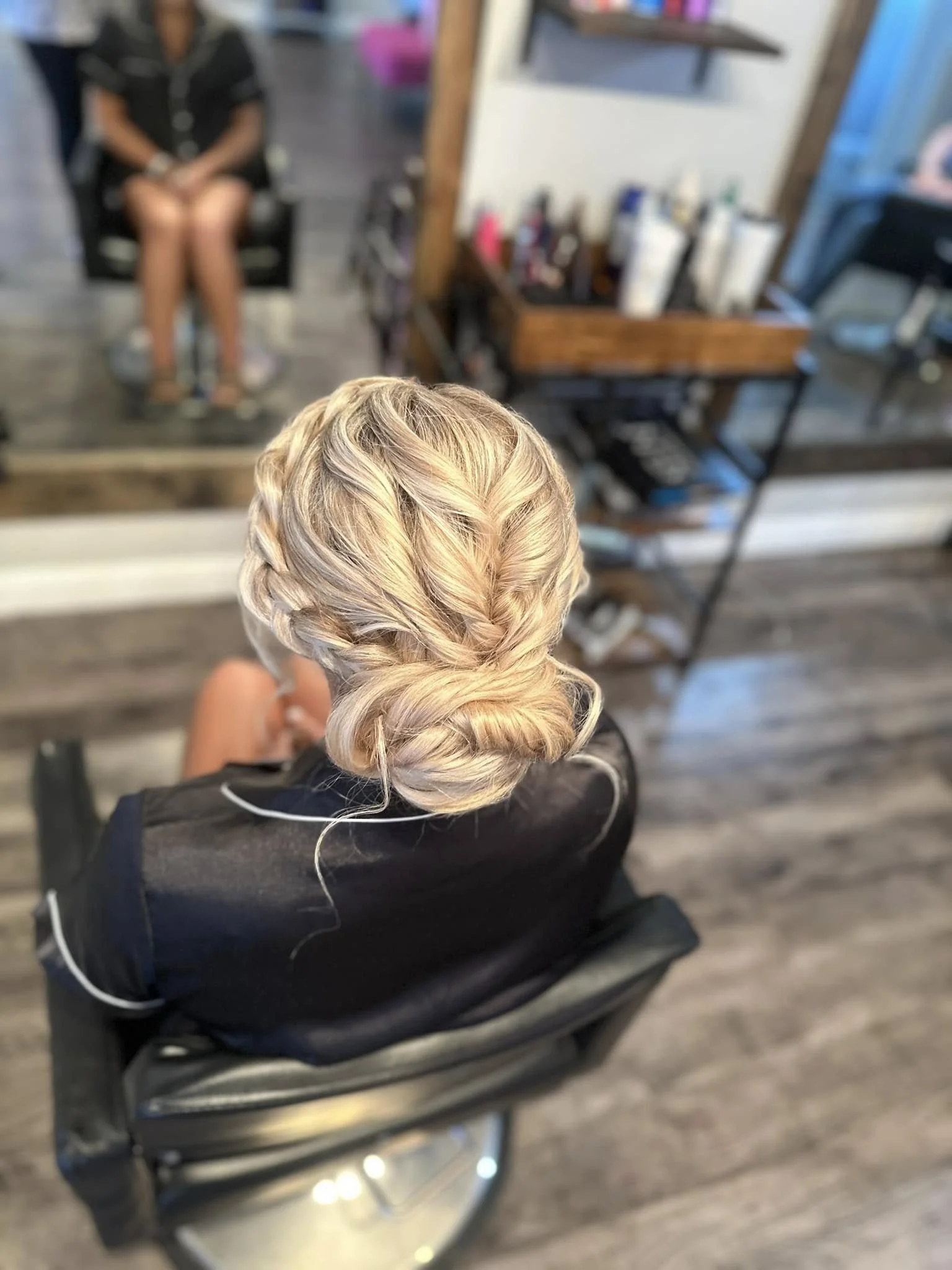A woman with blonde, braided and curled hair sitting in a salon chair, with her reflection visible in a mirror behind her showing another woman seated in a salon chair.